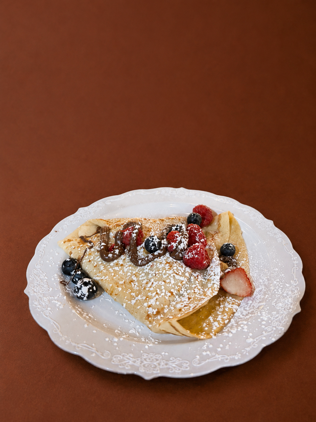Crepe filled with strawberries and blueberries, topped with chocolate syrup and powdered sugar on a decorative white plate.