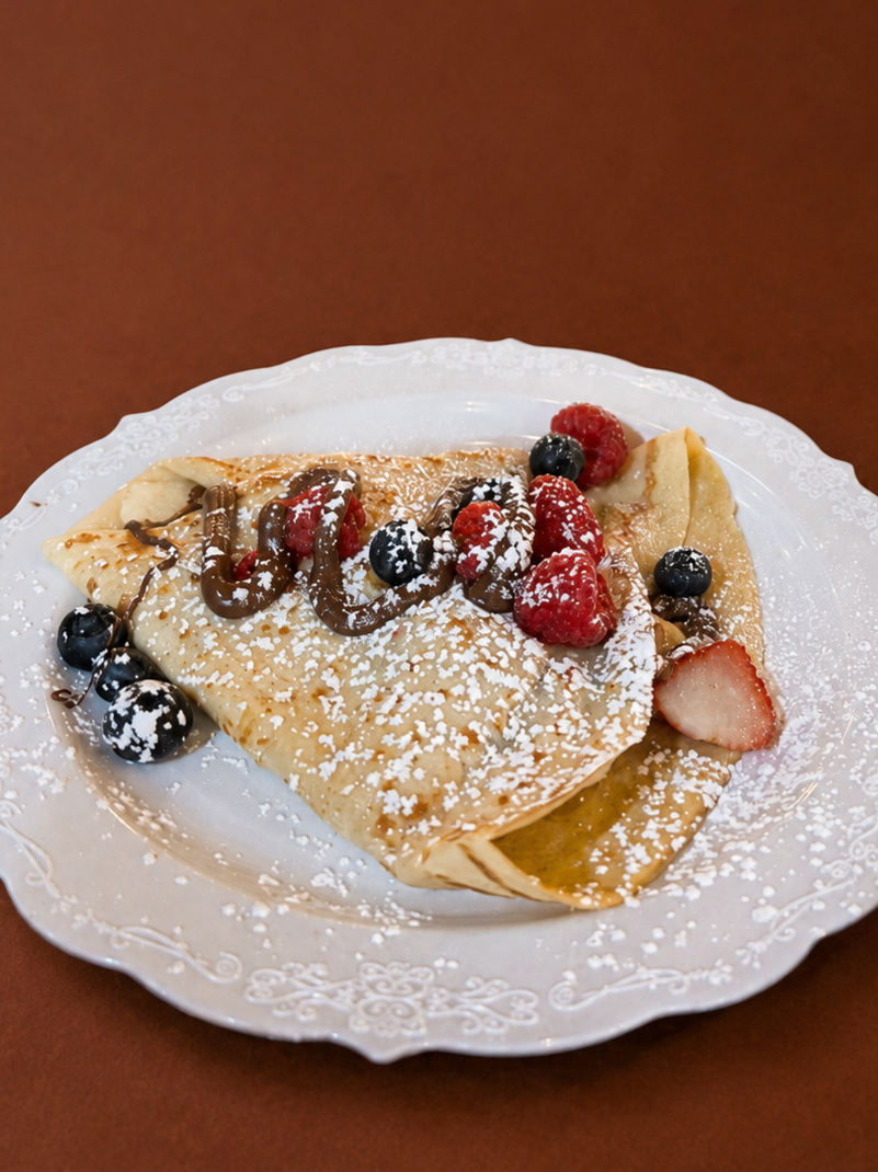 Crepe topped with mixed berries, chocolate sauce, and powdered sugar, served on a decorative white plate.