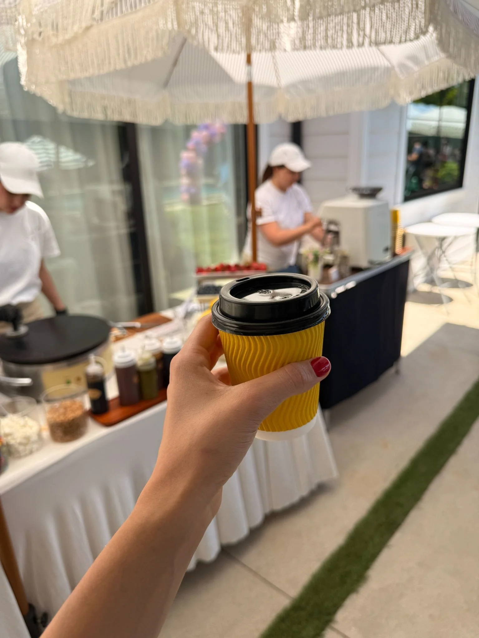 Person holding a yellow paper coffee cup with black lid in front of a coffee stand with two women in white shirts and hats preparing drinks.