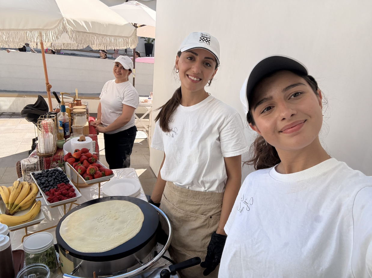 Three women at an outdoor fruit stand with a crepe in an electric pan, strawberries, blueberries, bananas, and other fruit on display.