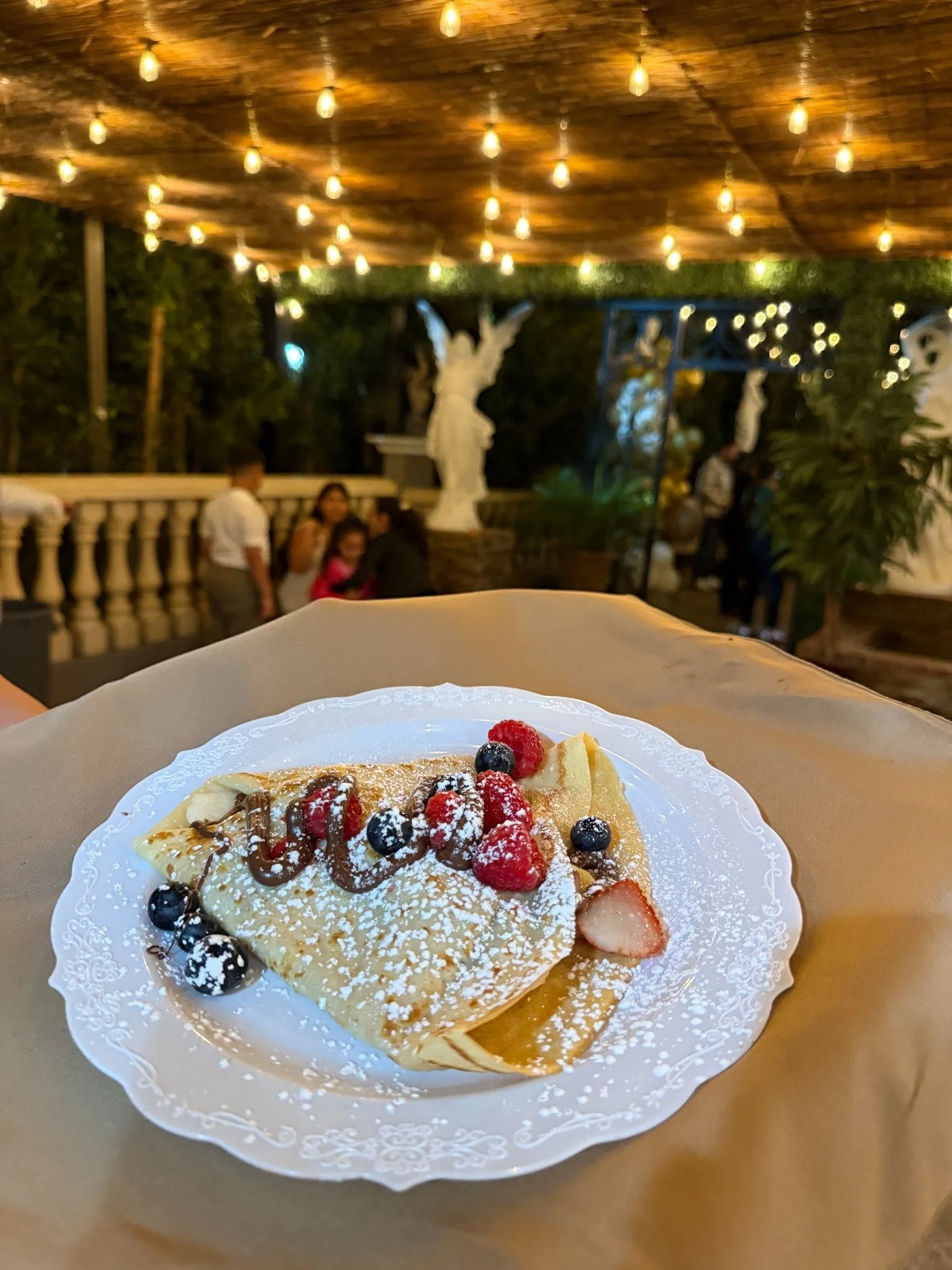 A plate of crepe topped with assorted berries, chocolate drizzle, and powdered sugar, set on a table in a restaurant or outdoor dining area with string lights and decorative statues in the background.