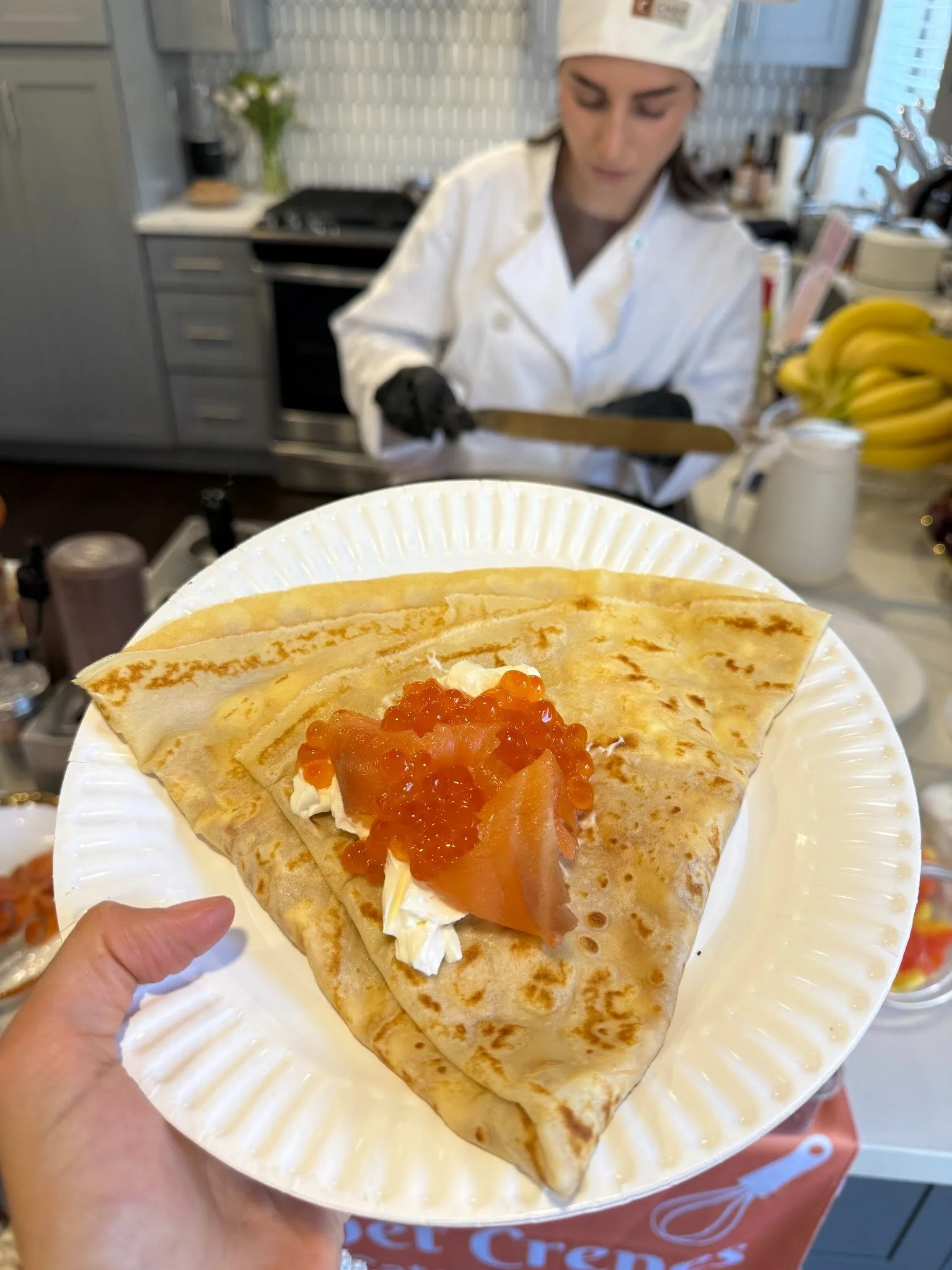 A close-up of a plate with two crepes topped with smoked salmon, crème fraîche, and red onion preserves, held by a person's hand in a kitchen. In the background, a person wearing a chef's hat and apron is working behind the counter.