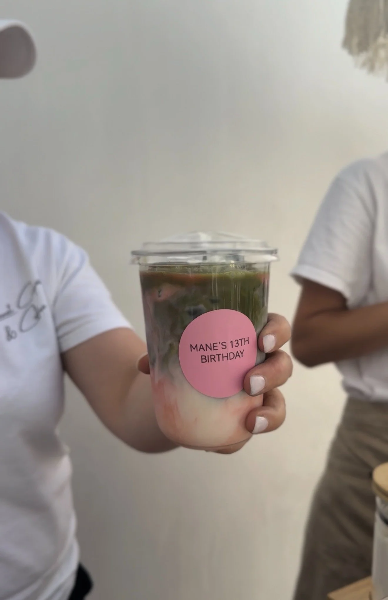 Person holding a cup with a pink sticker that reads "Mane's 13th Birthday" in a room with two other people, one wearing a white shirt and the other in beige pants.