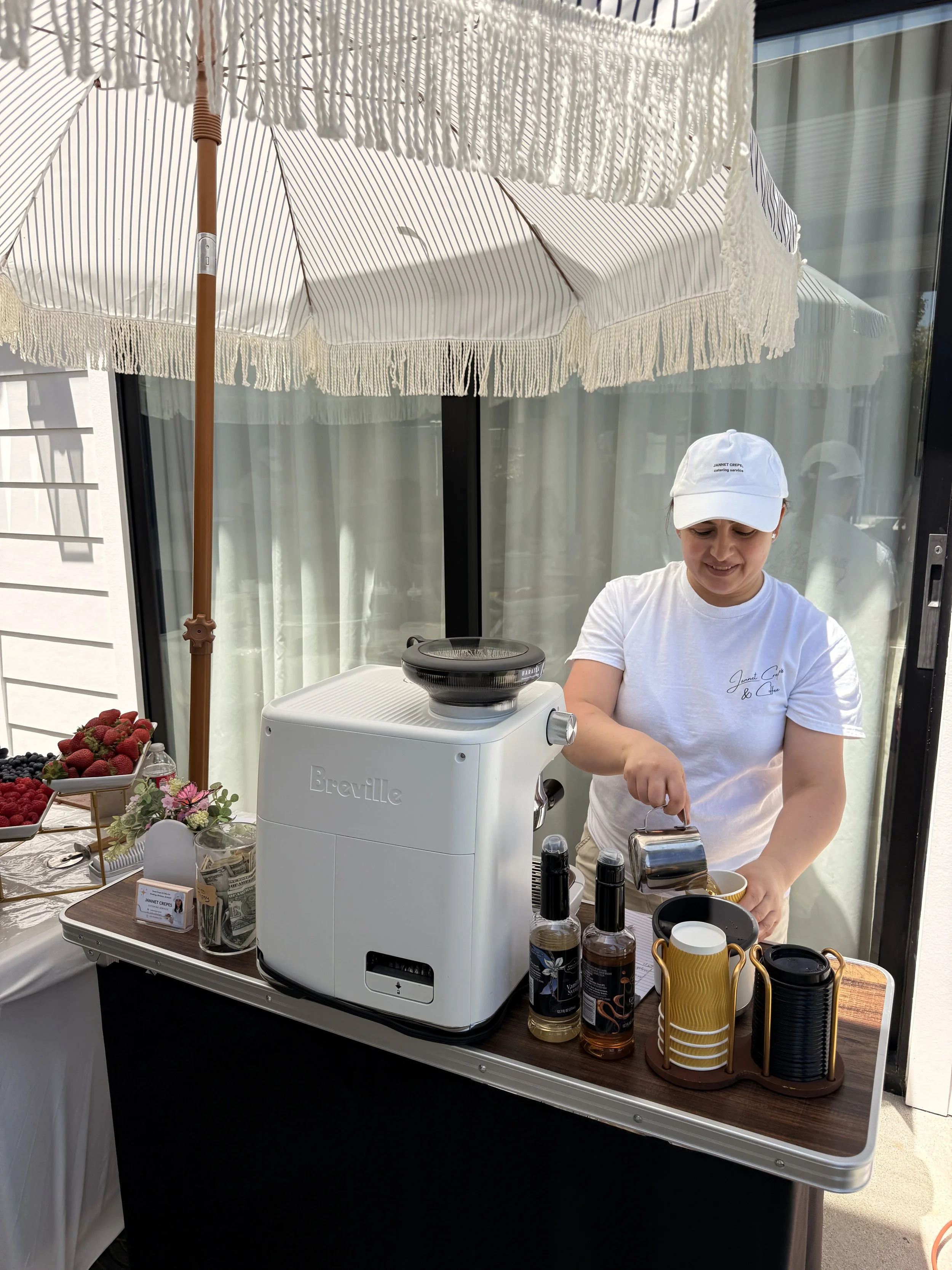 A woman wearing a white cap and t-shirt preparing coffee or a hot beverage at an outdoor market stall, with a Breville coffee machine, and various bottles and cups on the table. There are baskets of fresh berries and flowers, and a large white paraso