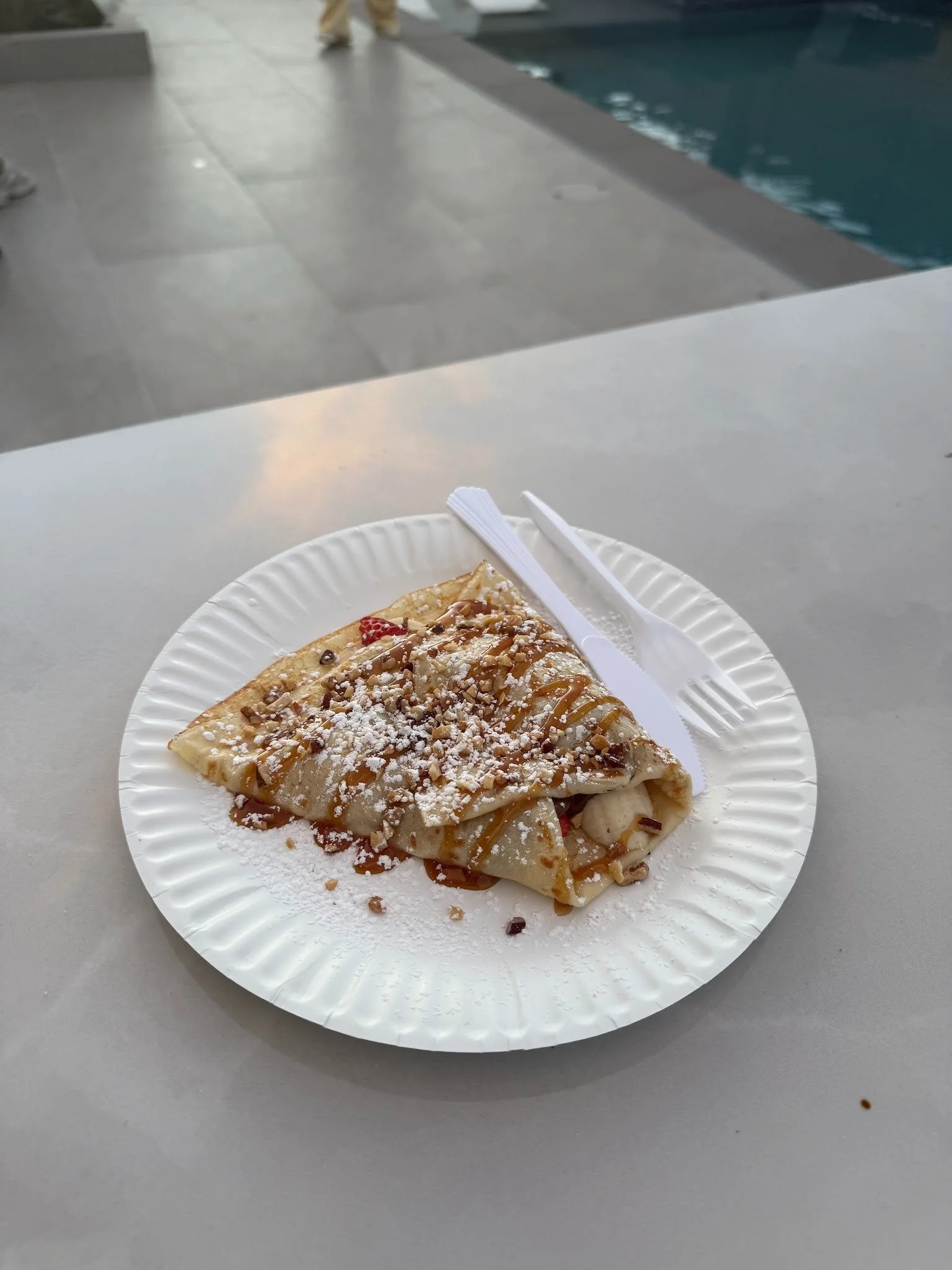A slice of crepe topped with powdered sugar, caramel drizzle, chopped nuts, and strawberries on a white paper plate, placed on a gray table near a swimming pool.