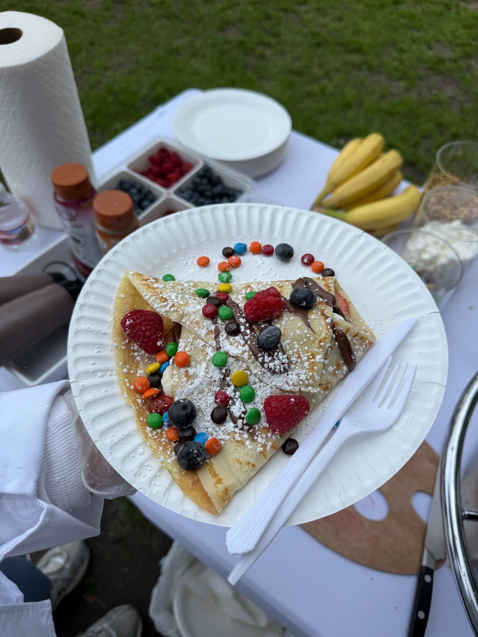 A paper plate with a slice of crepe pancake topped with berries, chocolate drizzle, colorful candies, and powdered sugar. In the background, there are bananas, small bowls of berries, whipped cream, and bottled condiments on a table outside.