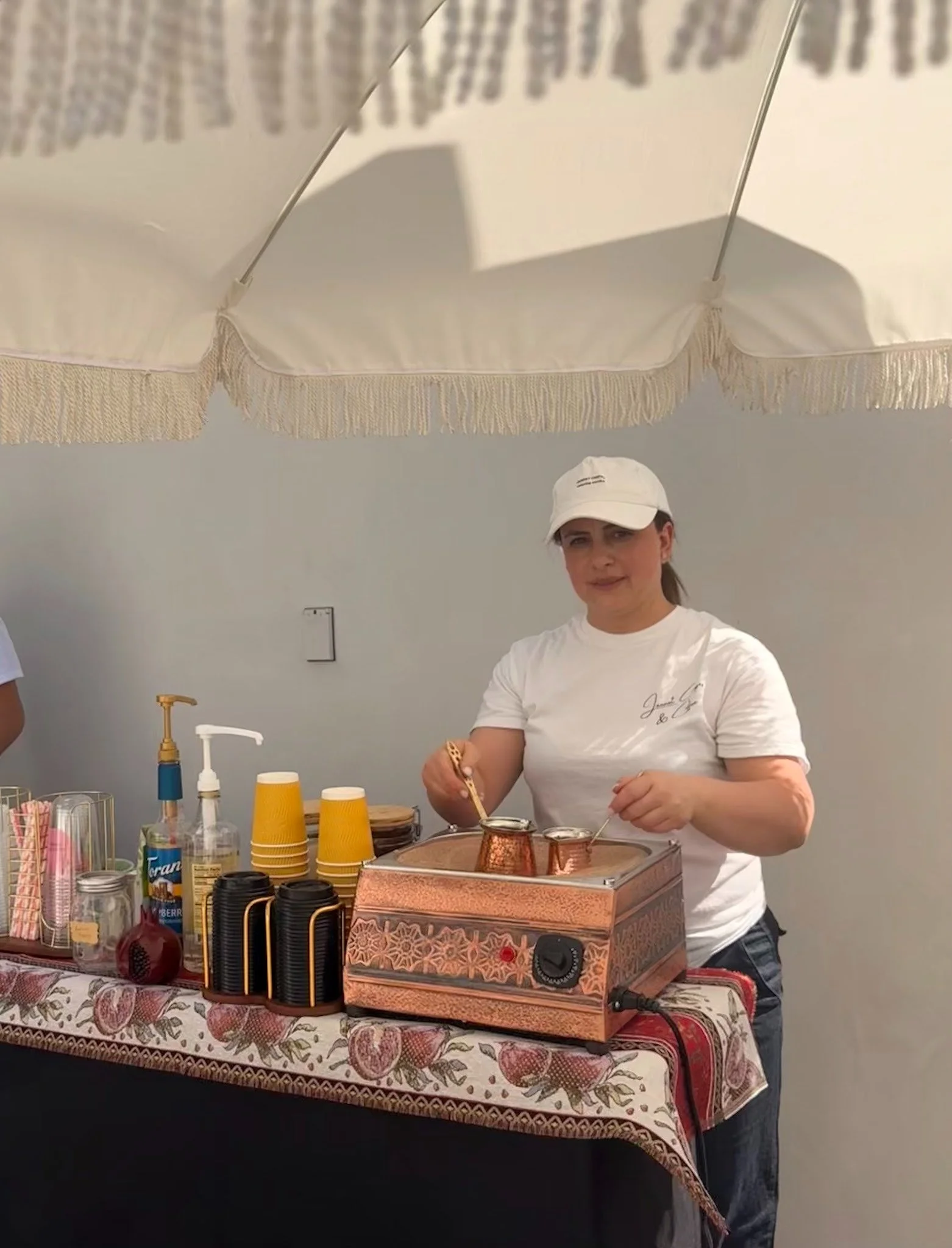 A woman wearing a white t-shirt and white baseball cap stands behind a table with an assortment of coffee cups, containers, and a copper-colored coffee machine, in a room with light-colored walls and a fringed white canopy overhead.