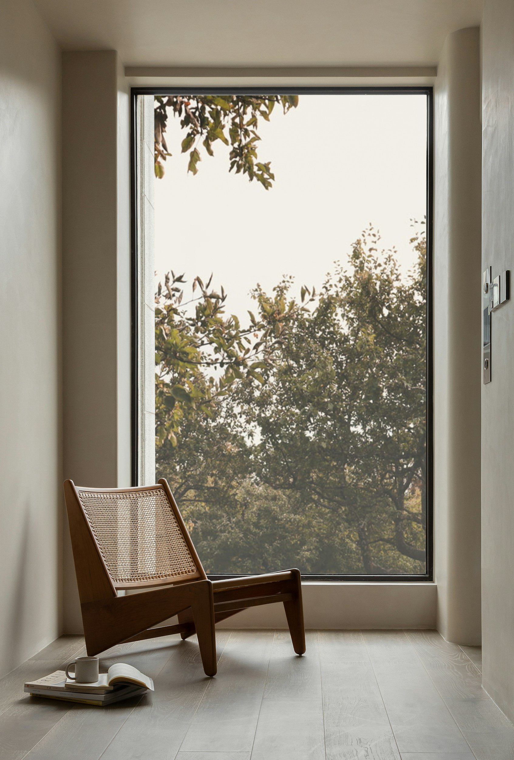 A wooden chair with a woven seat in front of a large window overlooking trees. On the floor, there are a few open books, a white mug, and a closed book.