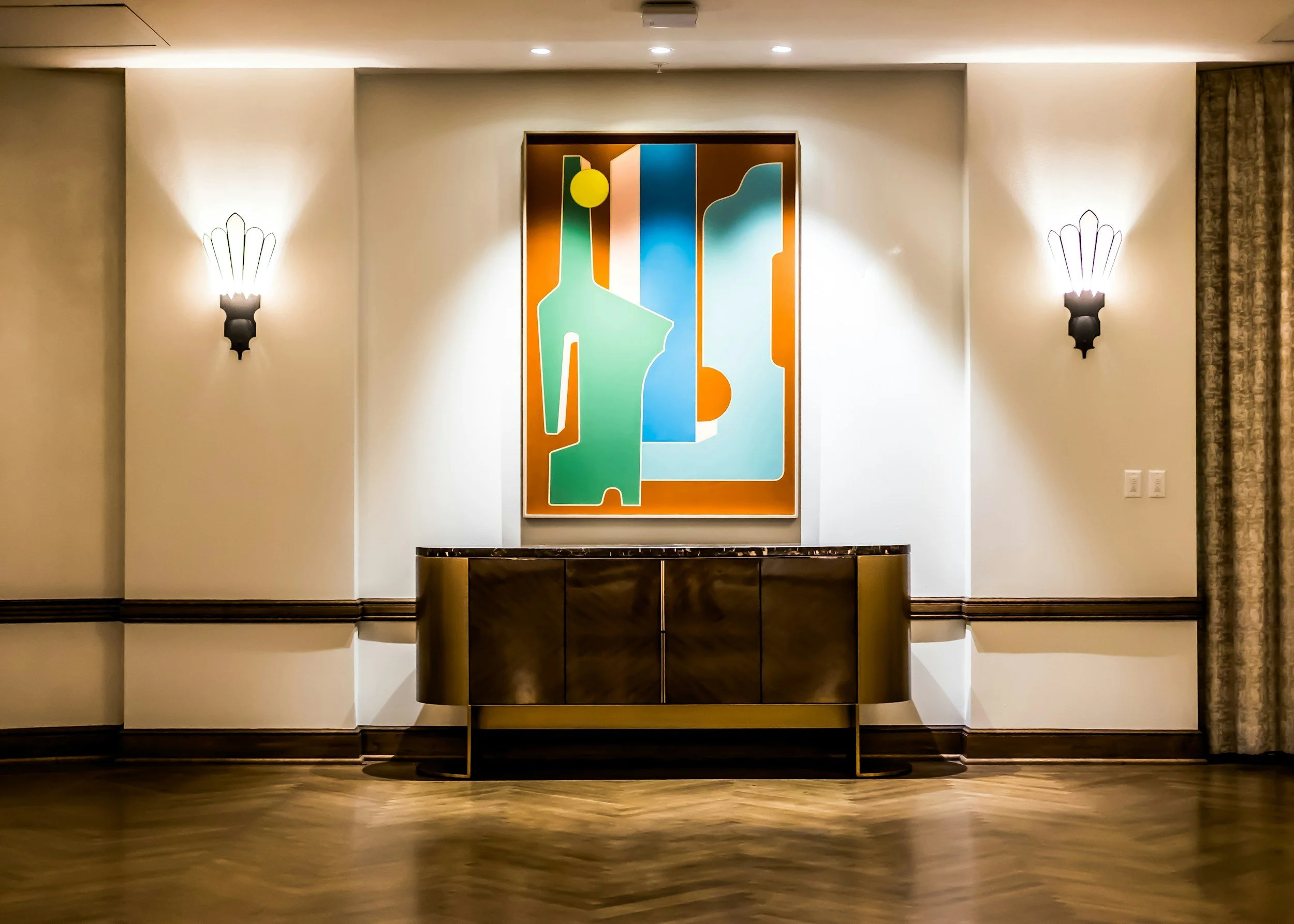 Decorative wall with two wall sconces and abstract art piece, a curved wooden console table with a marble top below the artwork, beige curtains partially visible on the right, and wood flooring.