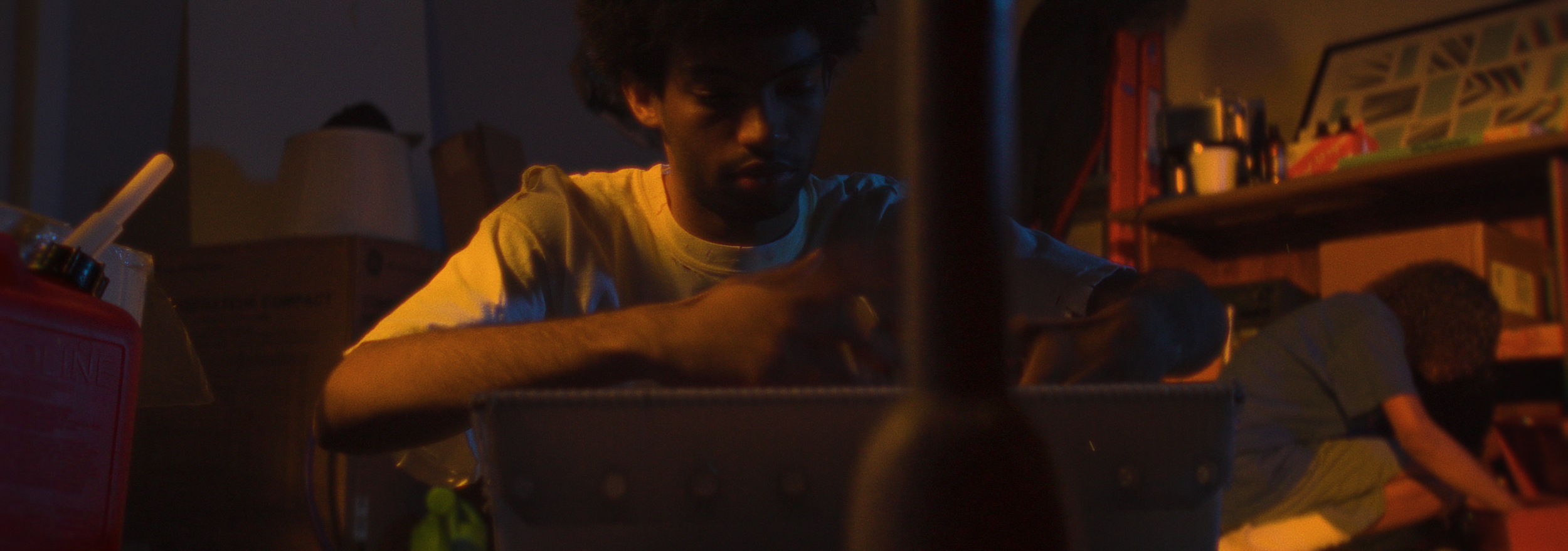 A young man with dark curly hair in a dimly lit room, focused on working on a project or task at a cluttered table with various objects including a red container, a lamp, and shelves with books and decor in the background.