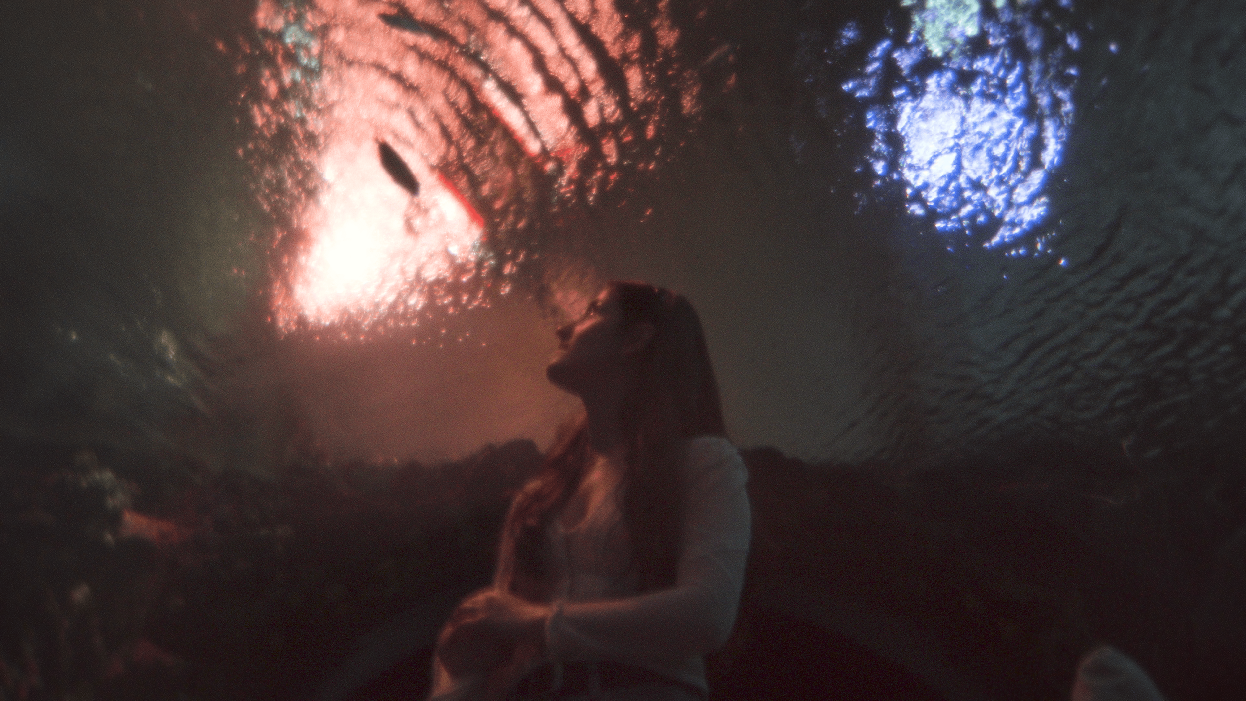 A person sitting underneath fireworks in the night sky, some of which are blue and others are red and white.