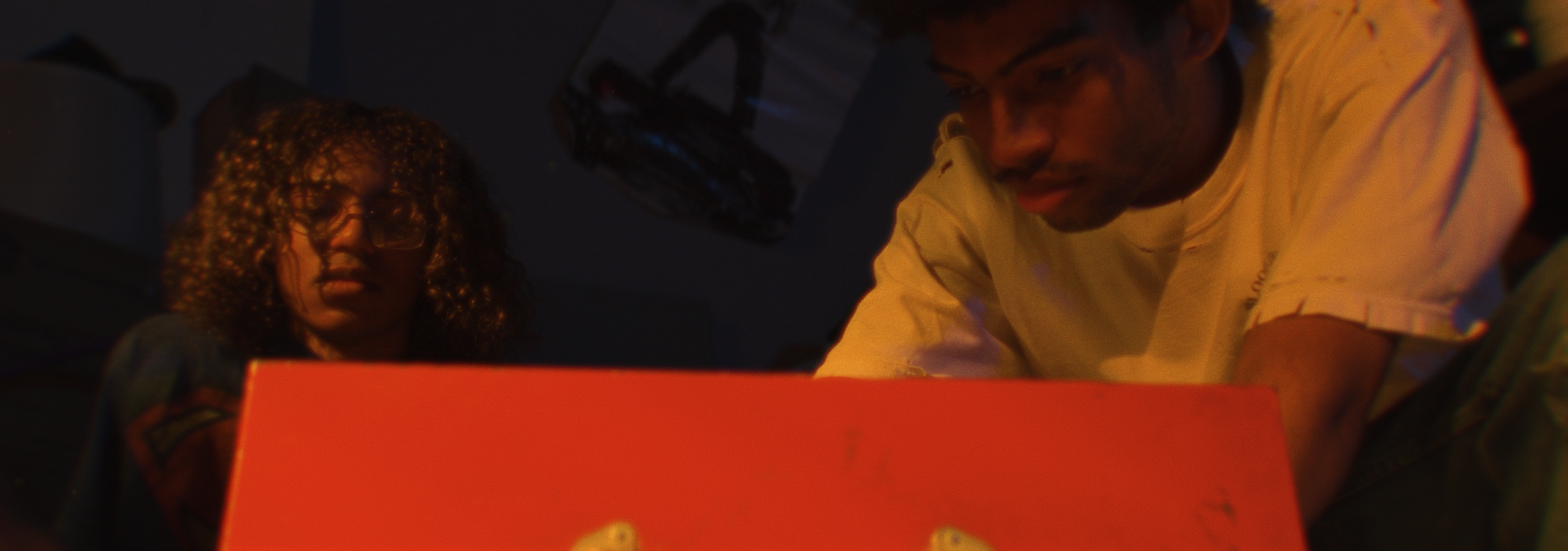 Two people, a man with curly hair and glasses and a man with short hair, looking at a bright orange screen or object in a dimly lit room.