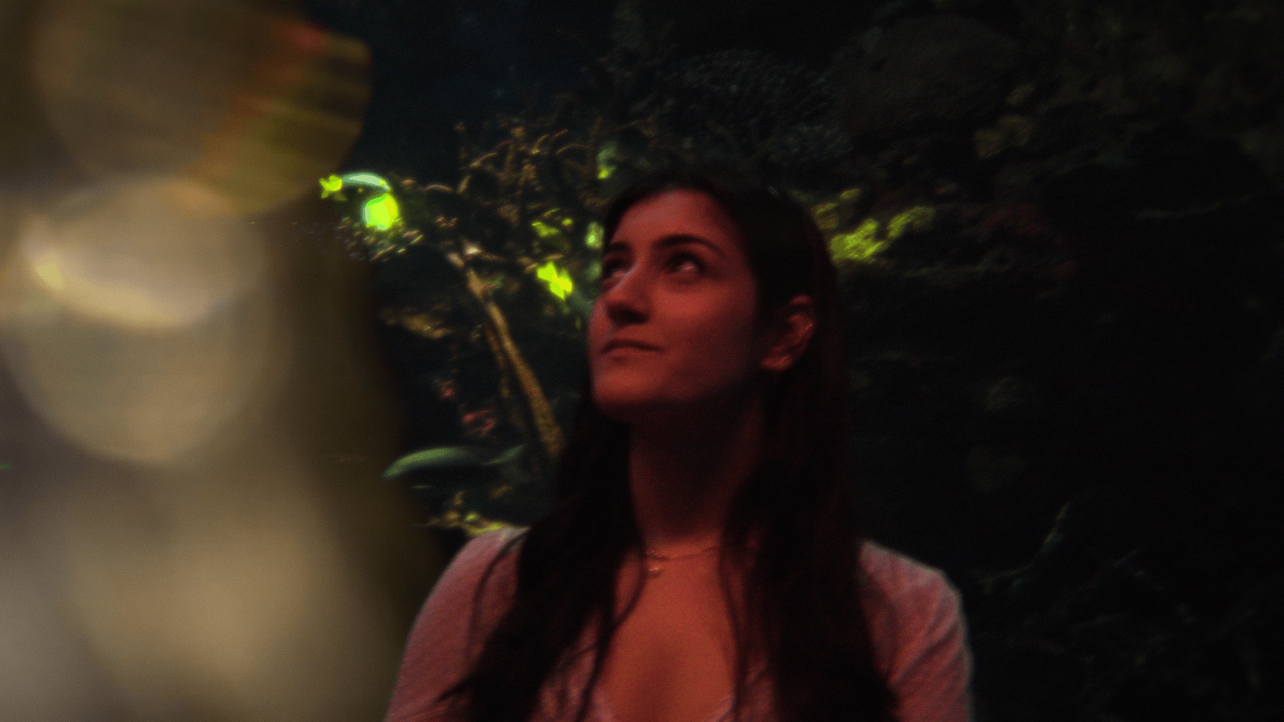 Close-up of a woman with long dark hair looking upward in a dark environment with greenish lighting and blurry foreground elements.
