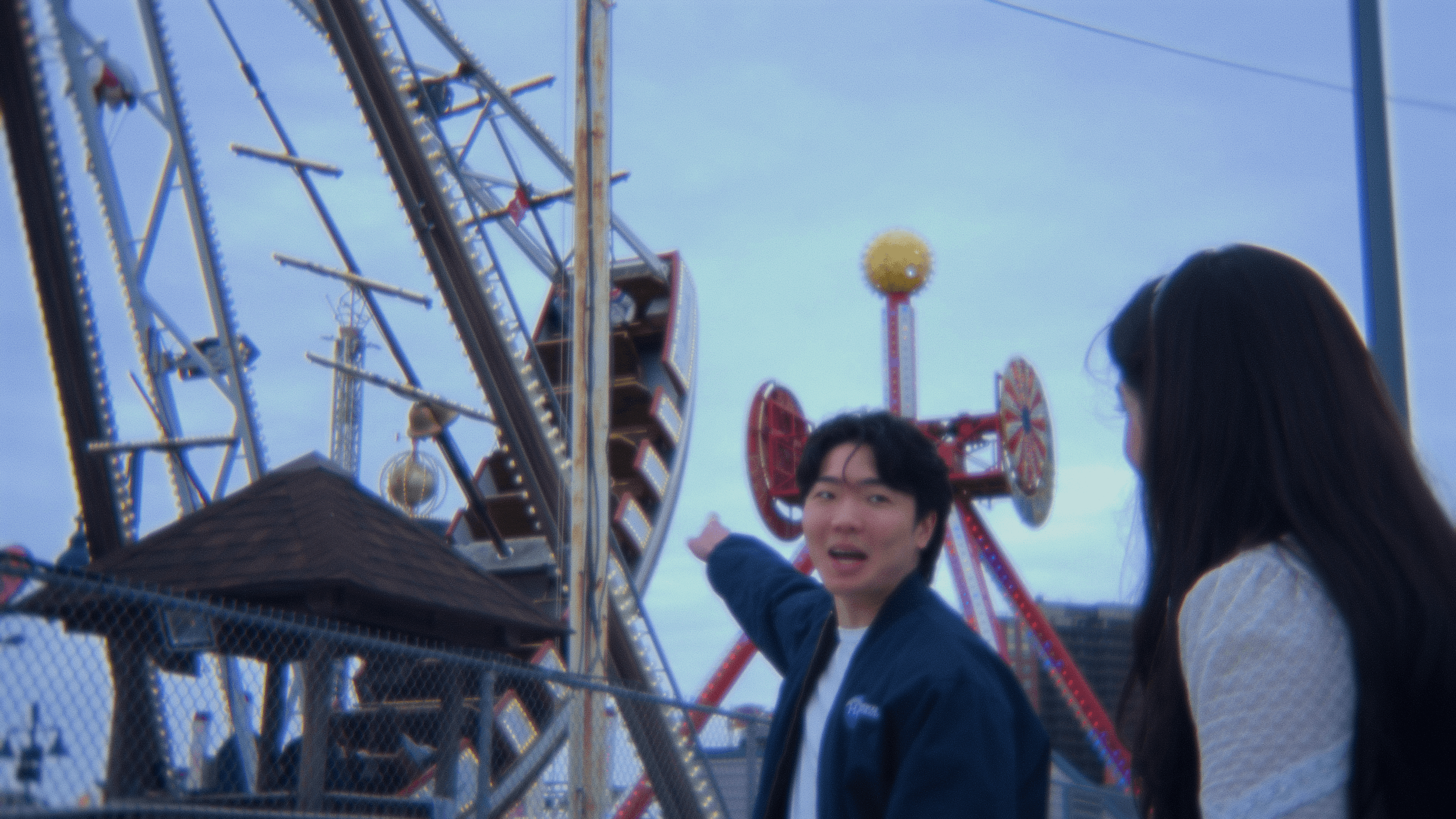 Two people, a young man and woman, are at a carnival or amusement park near a large Ferris wheel and other rides. The young man is pointing toward the rides and appears to be talking, while the woman is looking in the same direction. The sky is partl