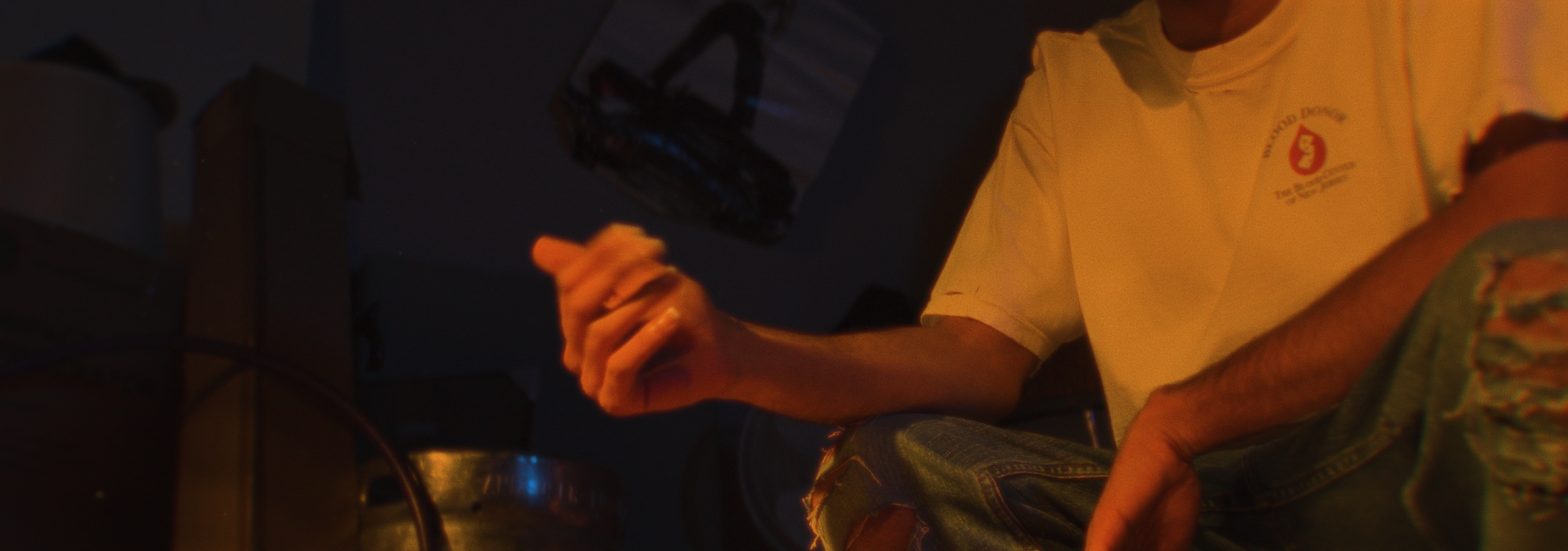 A person sitting in a dark room, wearing a yellow T-shirt and camo shorts, holding out their hand.