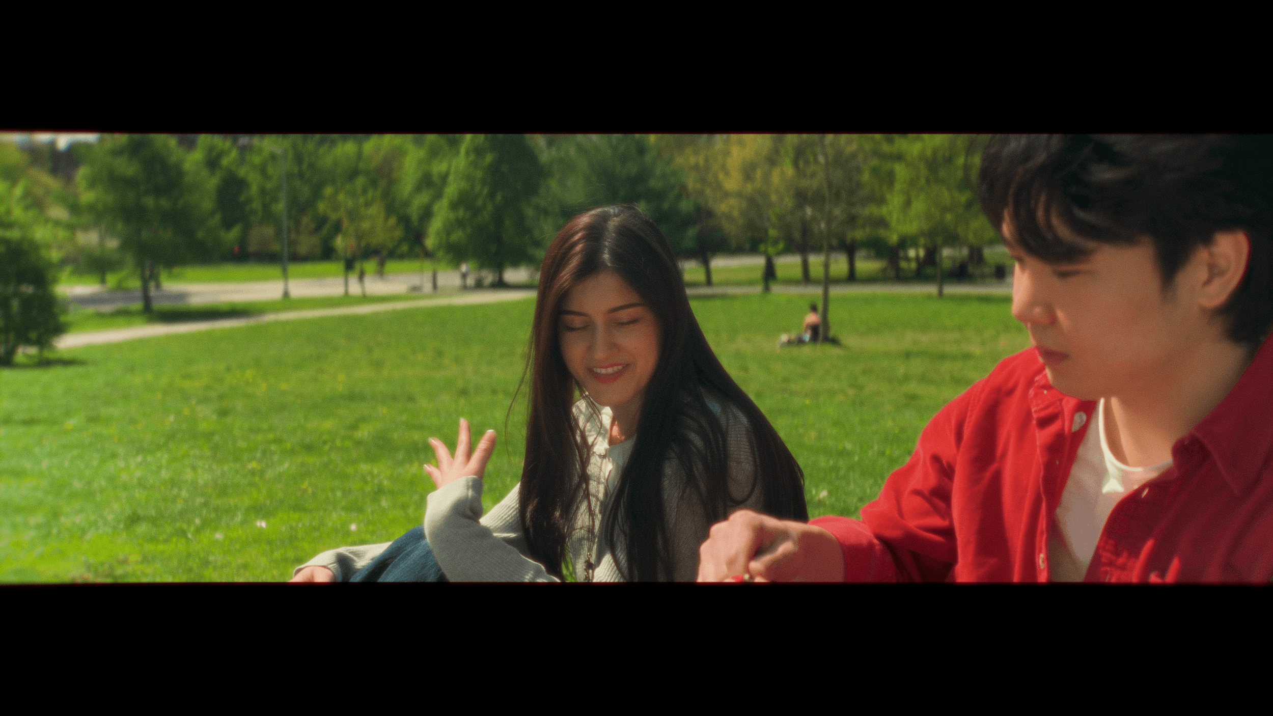 Two young people, a woman with long dark hair and a man with short dark hair, sitting on grass in a park, smiling and looking at a phone. Green trees and a walking person in the background.
