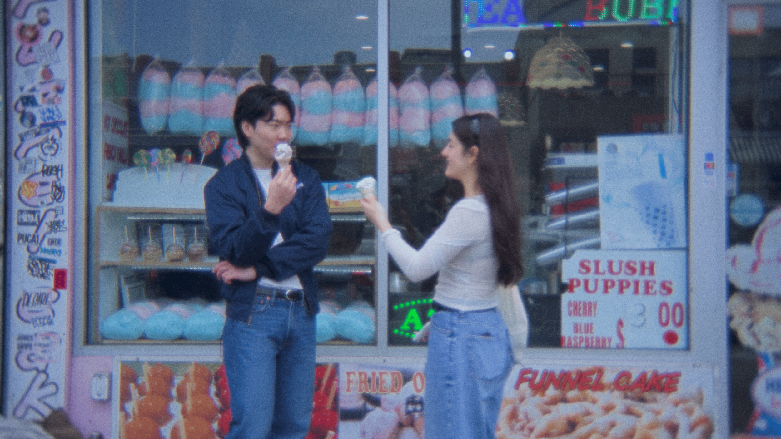 A young man and woman are standing outside a colorful candy and ice cream shop. The man is holding an ice cream cone and looking at the woman, who is smiling and reaching out to take it. The shop has bright signage and displays of marshmallow candies