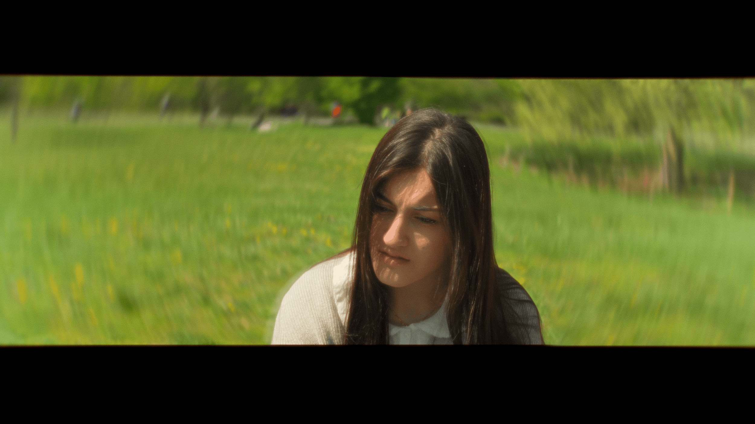 Young woman with long dark hair sitting outdoors on grass in a park with green trees in the background.