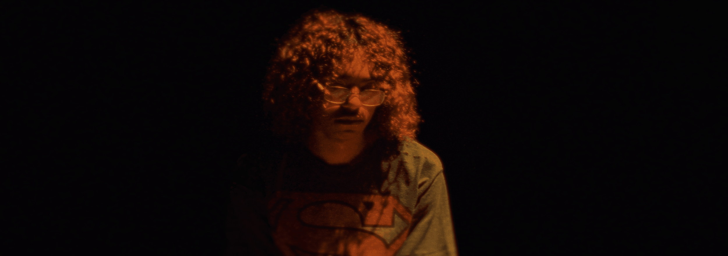 A person with curly hair, glasses, and a Superman logo shirt, illuminated by a warm orange light against a dark background.