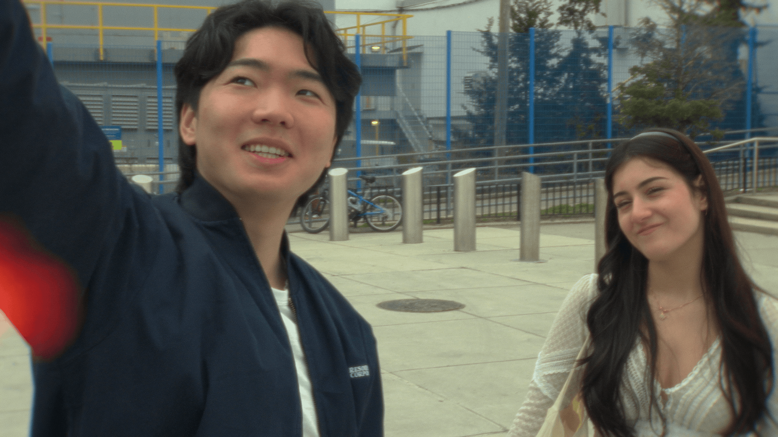 Two young women standing outside near a sidewalk with a blue fence and yellow railings in the background. One woman has dark hair and is smiling, while the other has darker hair and is smiling softly.