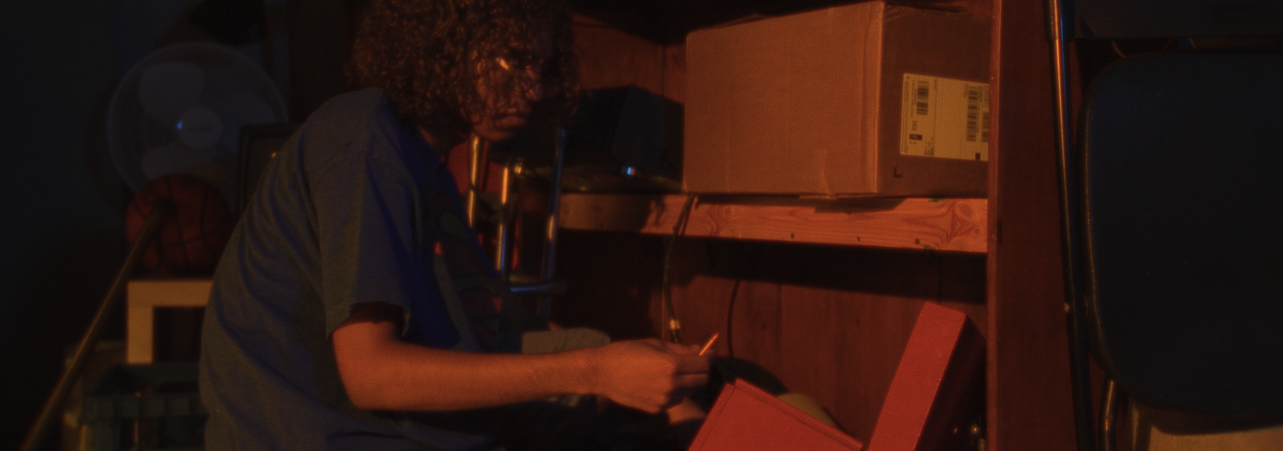 A young person with curly hair, sitting at a table in a dimly lit room, holding a bullet, with a cardboard box and a fan in the background.