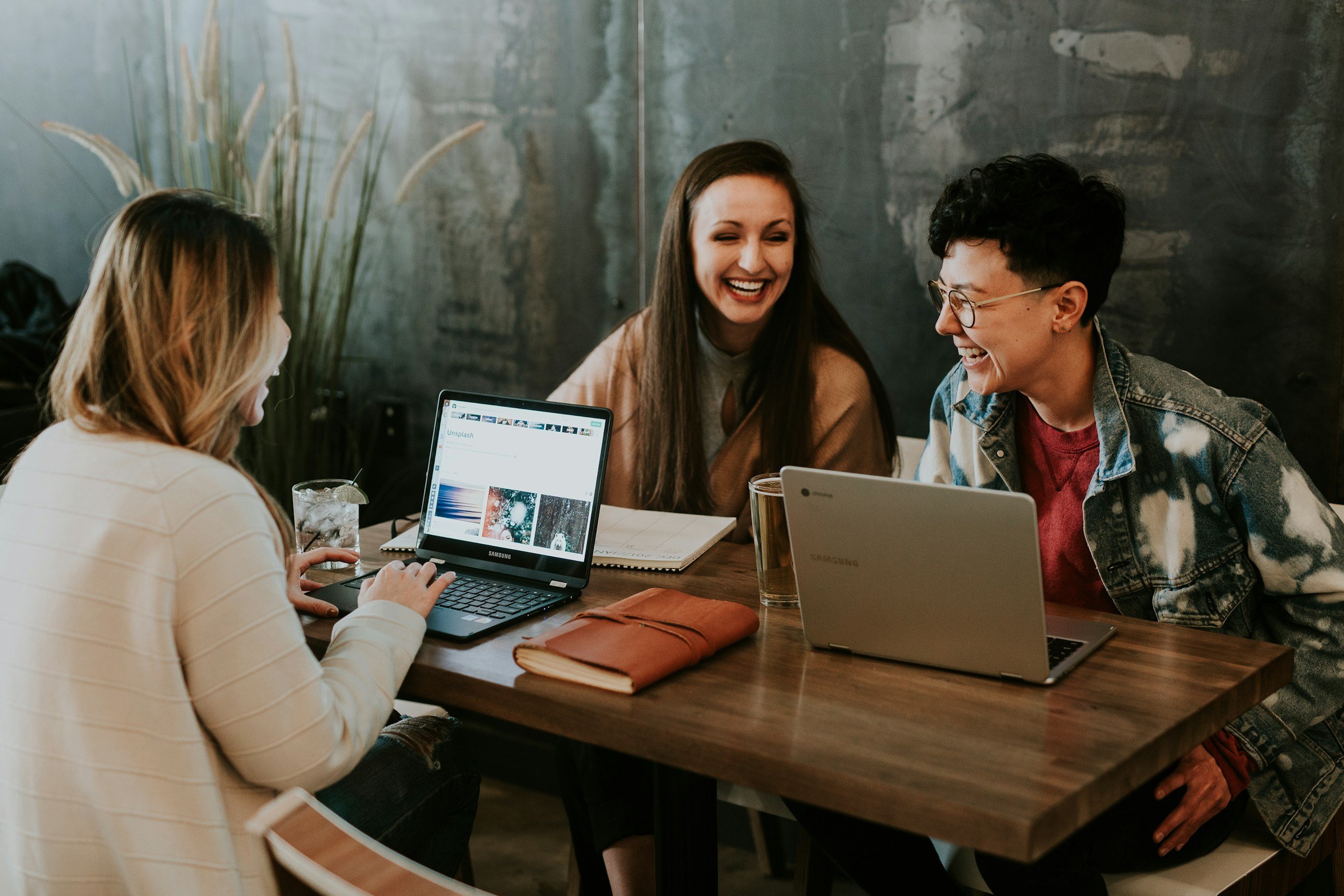 Four women sitting at a wooden table in a cafe, working on laptops and smiling, with drinks and notebooks on the table. One woman is wearing glasses and a denim jacket, all appear to be having a friendly discussion.