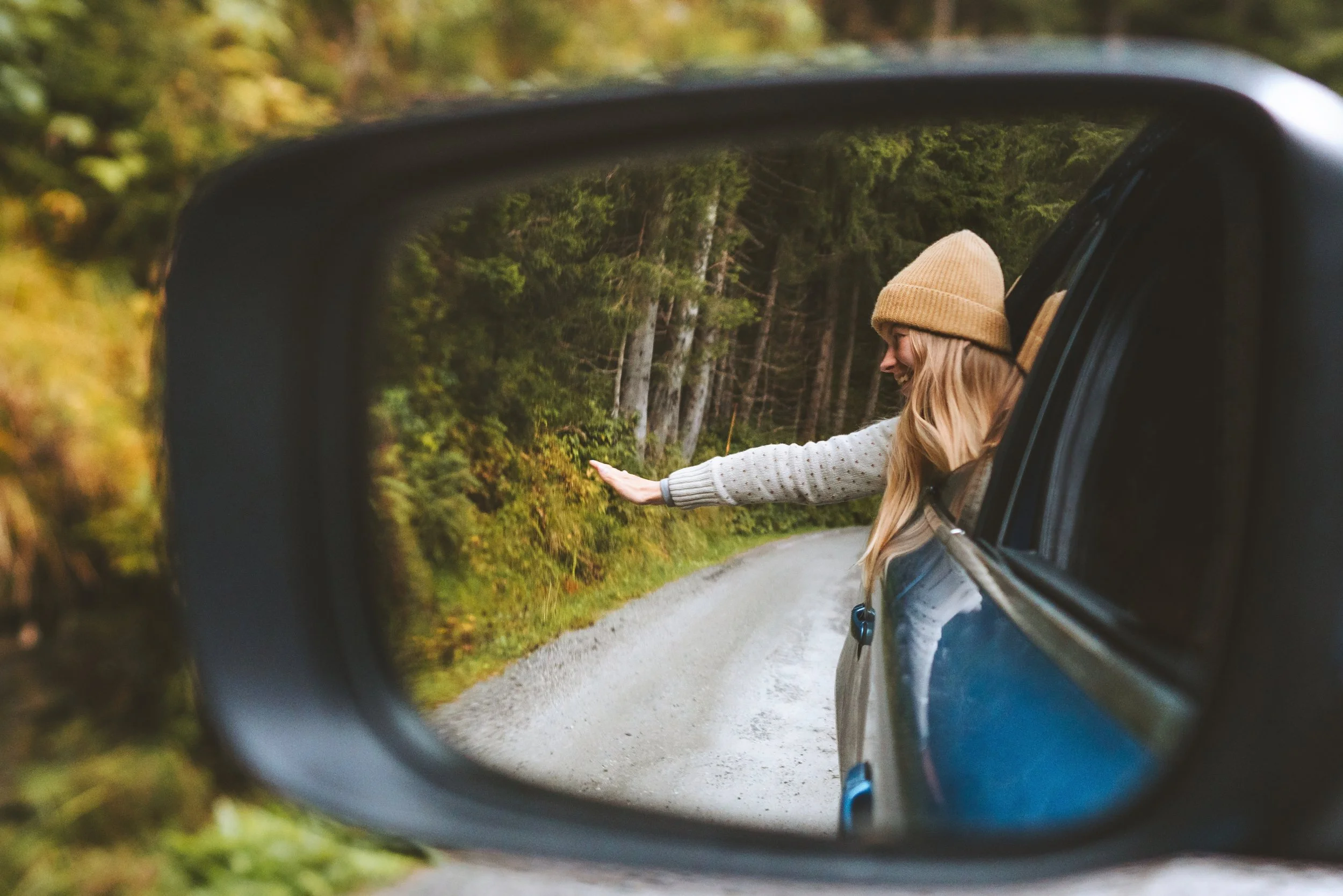 Side view mirror of a blue car showing a woman with blonde hair, a beige beanie, and a gray sweater extending her arm out and smiling, with a forest and a gravel road in the background.