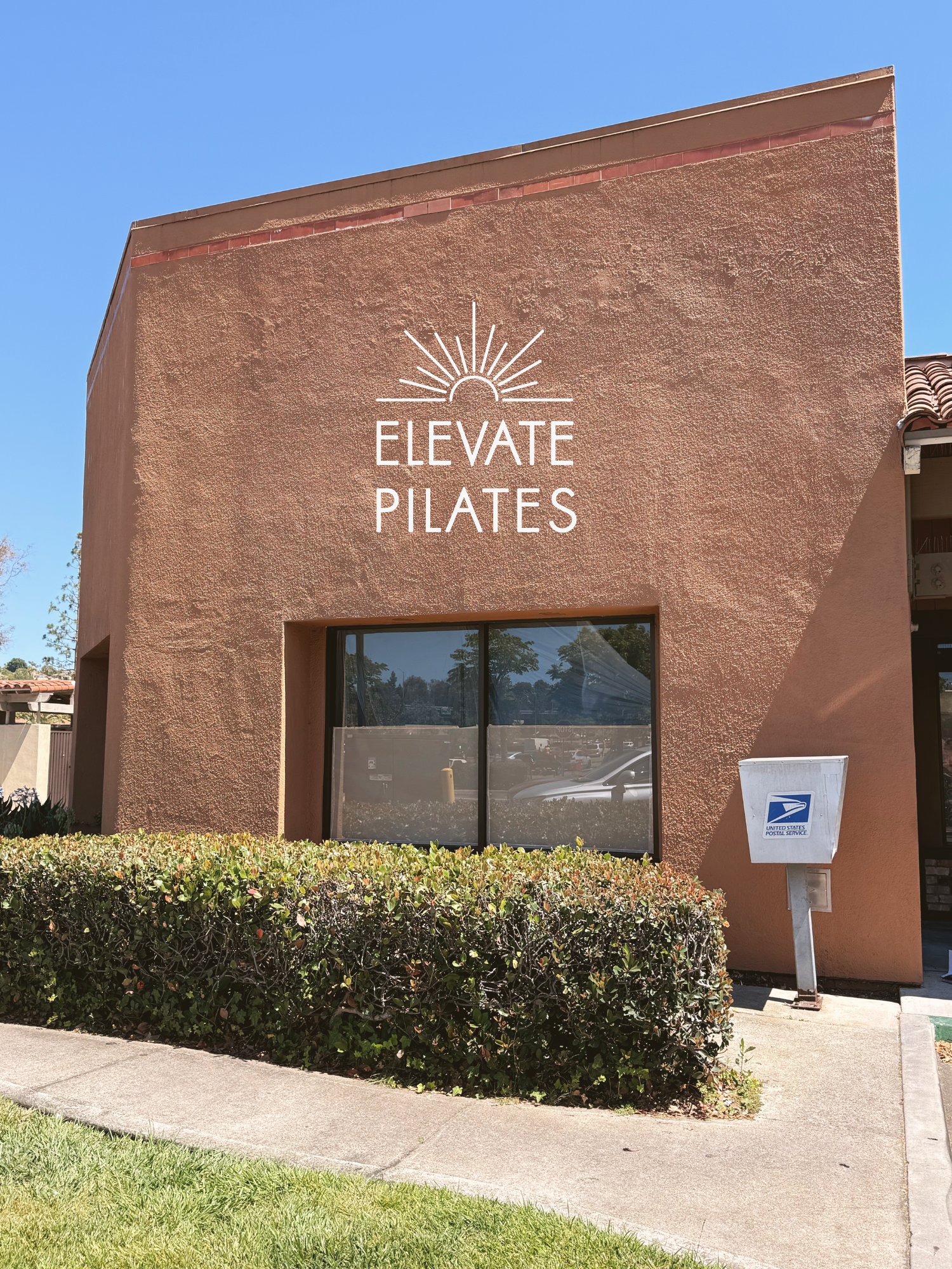 Exterior of a building with a sign that reads 'Elevate Pilates' and a sunburst logo. There is a hedge, sidewalk, and a United States Postal Service mailbox in front.