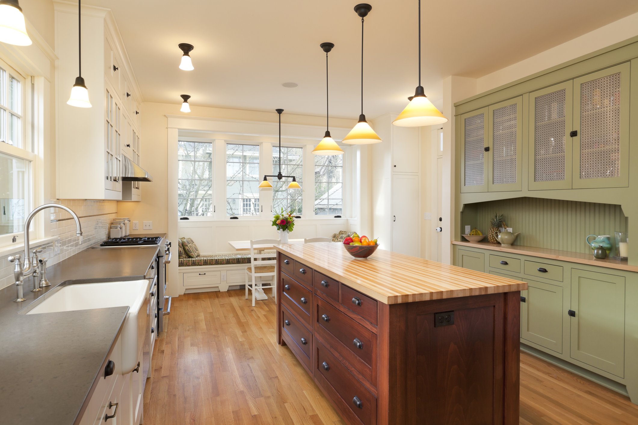 Bright, modern kitchen with white cabinetry, a large wooden kitchen island, green cabinets, and pendant lights hanging from the ceiling. Large windows provide natural light, with a seating nook by the window.
