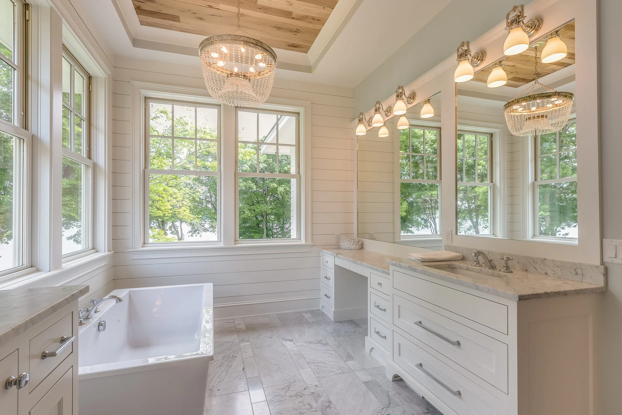 Bright bathroom with large windows, white cabinetry, marble countertops, a bathtub, and a mirror with lighting. Wooden ceiling panel and chandelier also visible.