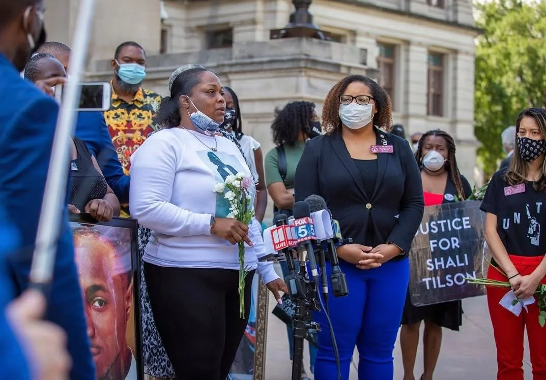 Group of people protesting outdoors, some holding flowers and signs, with microphones set up for a speaker, in front of a historic building.