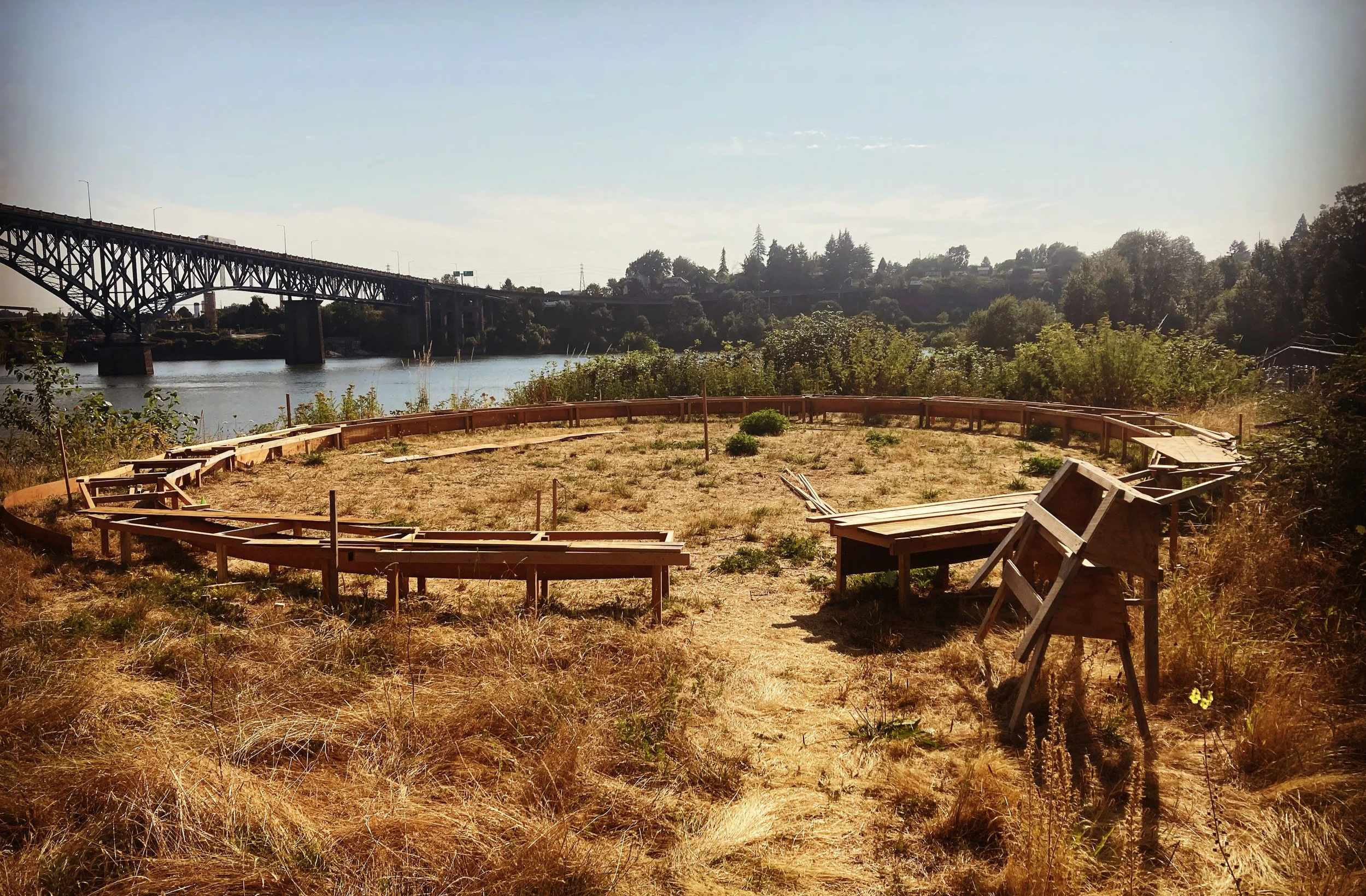An empty circular wooden bench under construction in a field of dry grass with bright sun on the banks of the Willamette River - a piece by sculptor Philip Krohn with a sound installation by Seth Nehil.