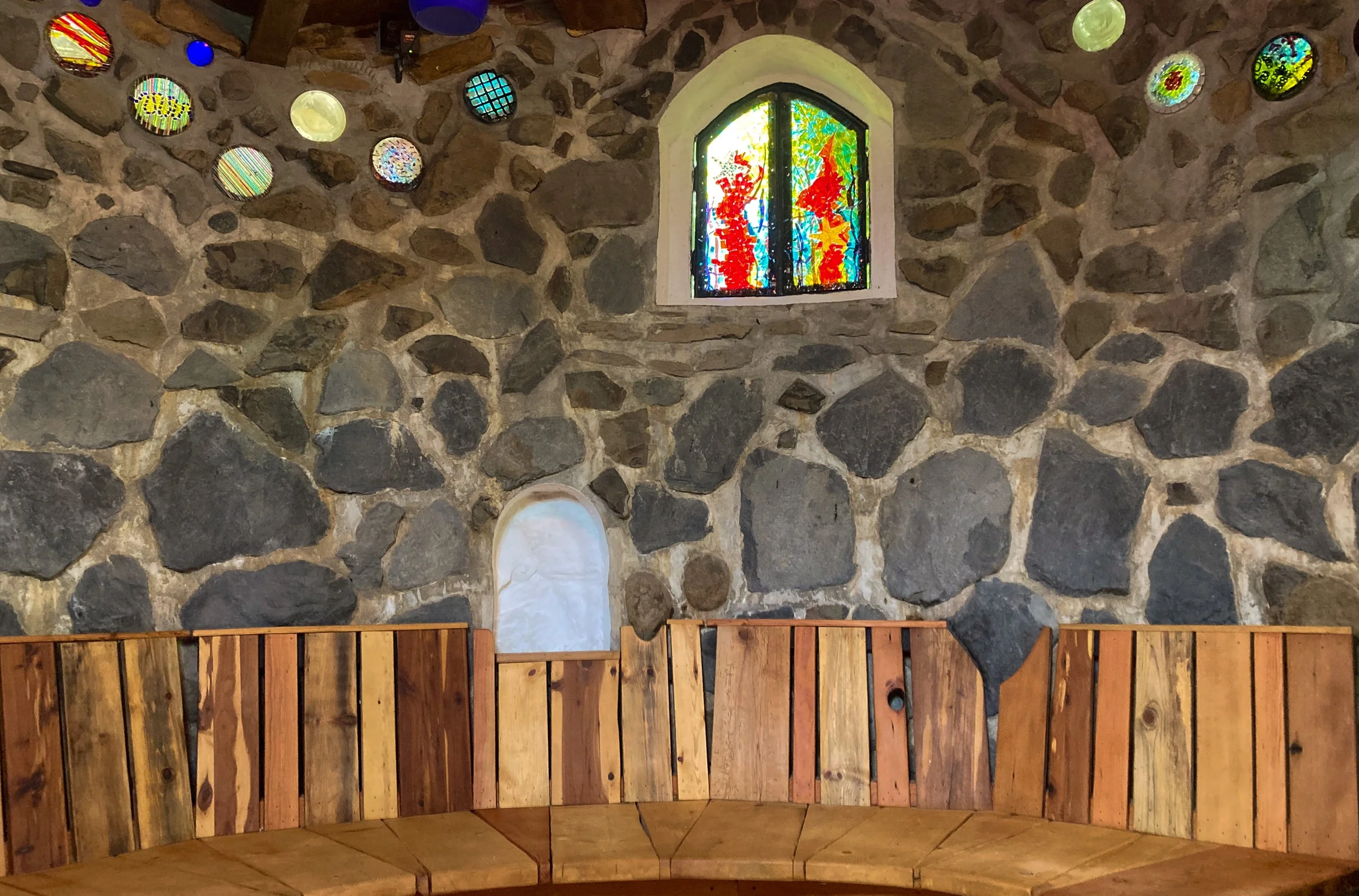 A long, handmade wooden bench and colorful fused-glass arched window.