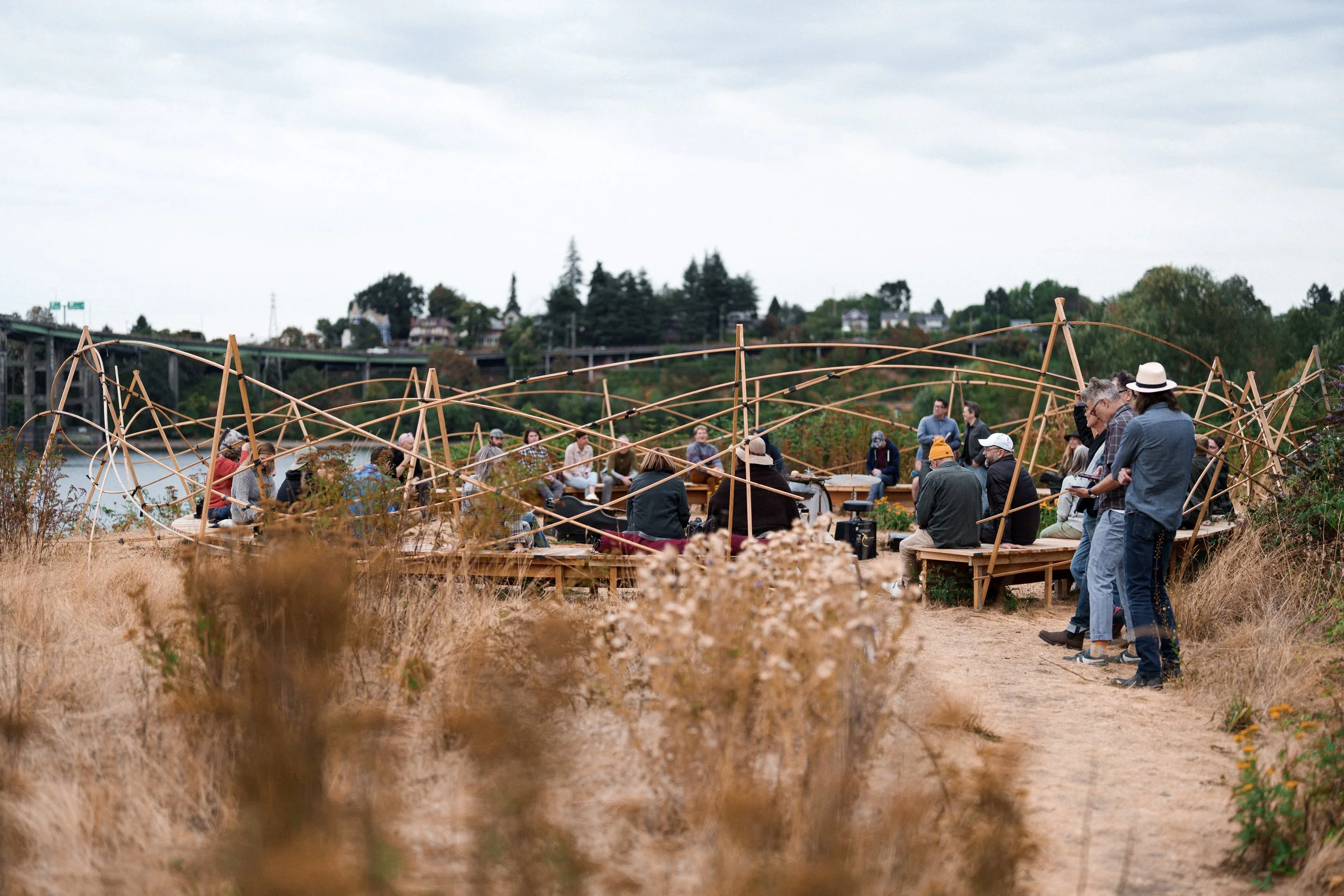 A group of people sit on a circular wooden bench in a field of dry grass on the banks of the Willamette River - a piece by sculptor Philip Krohn with a sound installation by Seth Nehil.