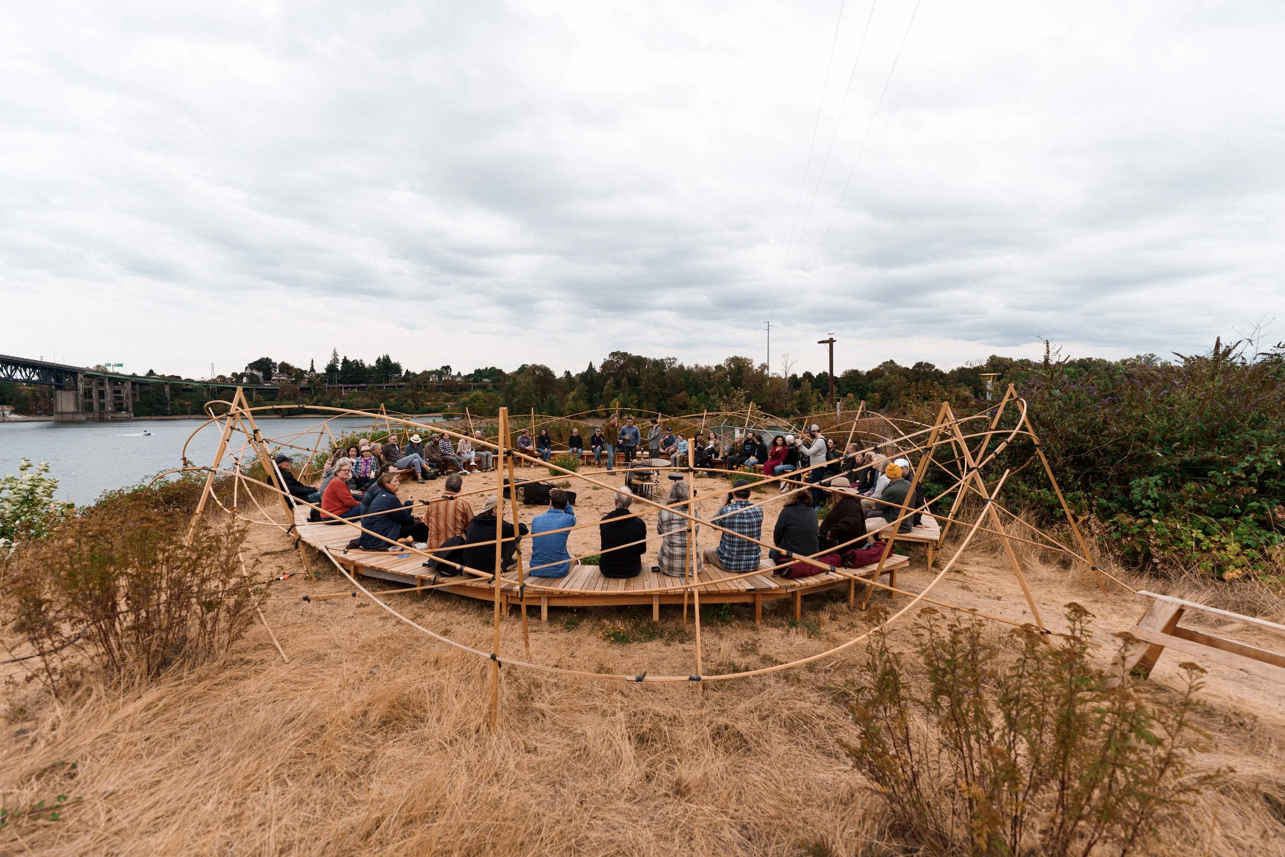 A group of people sit on a circular wooden bench in a field of dry grass on the banks of the Willamette River - a piece by sculptor Philip Krohn with a sound installation by Seth Nehil.