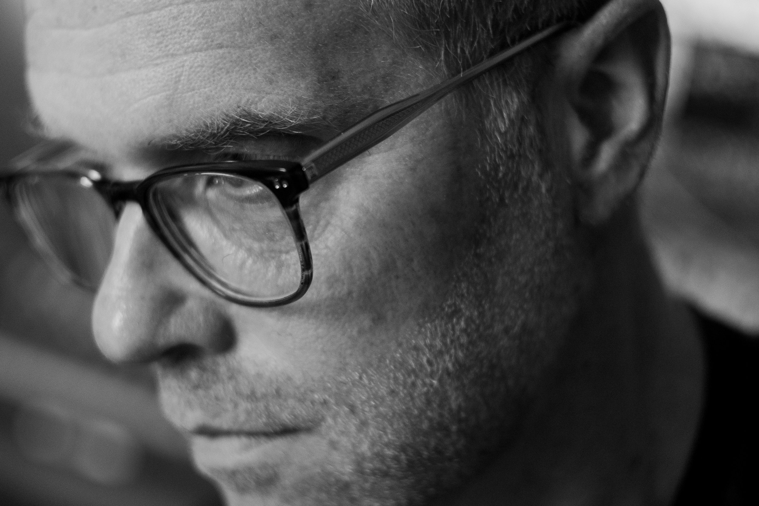 Close-up profile of a person's face wearing glasses, with a focus on the forehead, glasses, and a partial view of the ear in black and white.