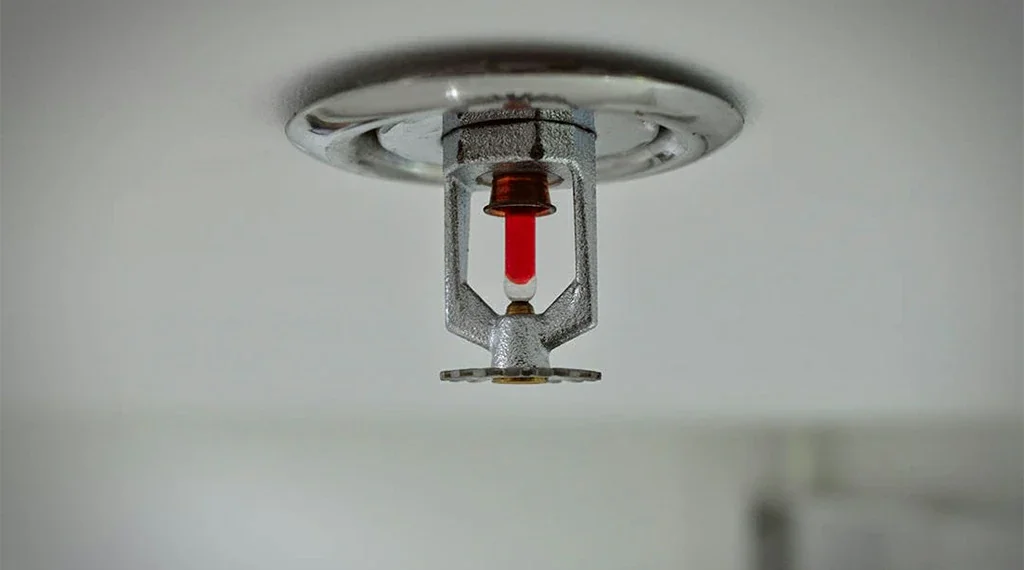 Fire alarm sprinkler head with red indicator on a ceiling.
