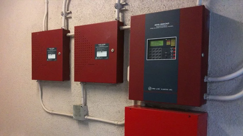 Four red fire alarm control panels mounted on a beige wall, connected by white conduit pipes.
