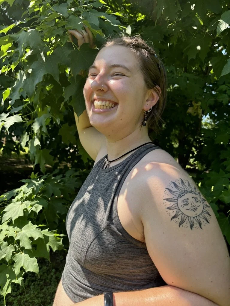 A young person with a tattoo of a sun with a smiling face on her upper arm, smiling and standing outdoors amidst green leaves and trees.