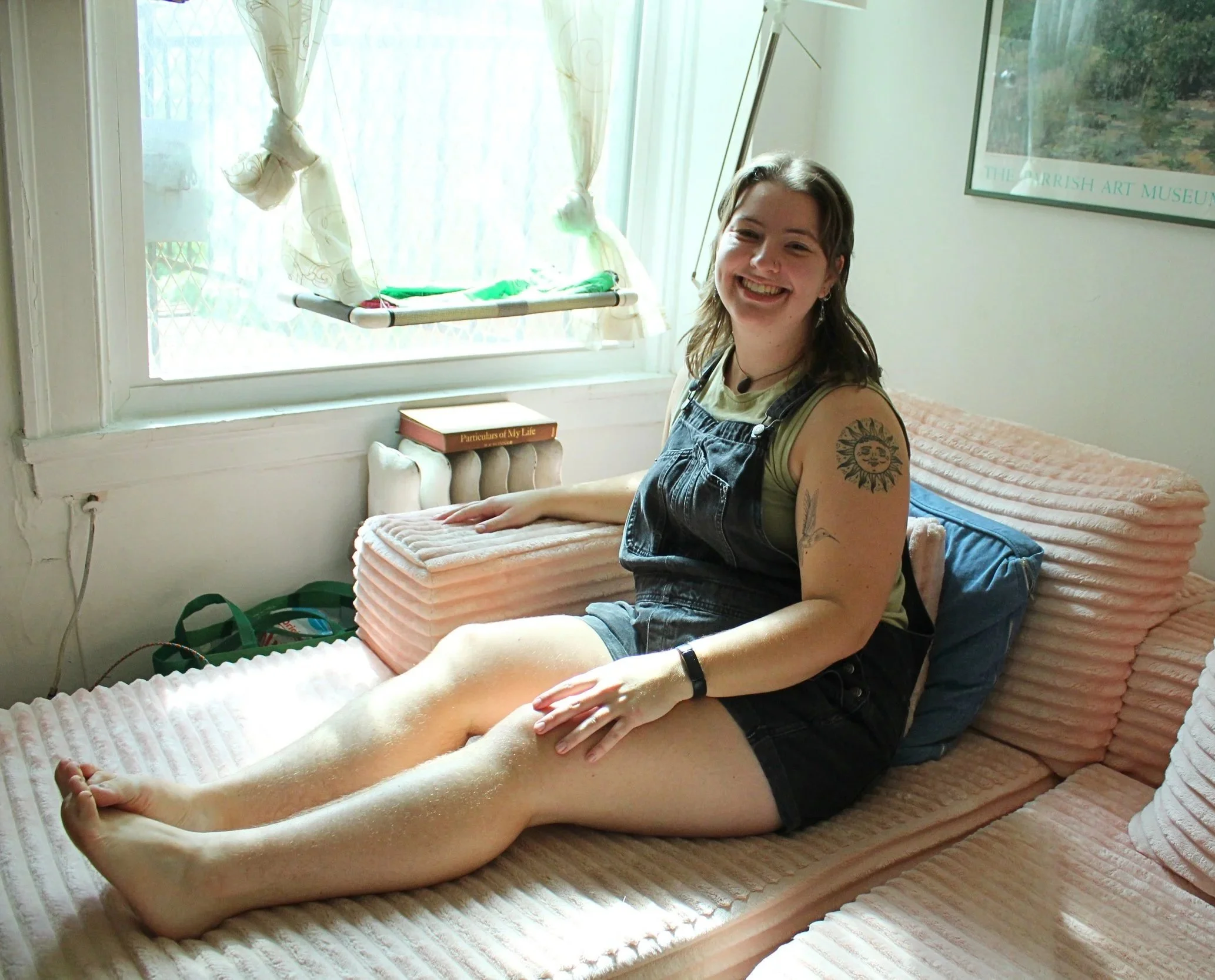 Home organizer Emilie smiling, sitting on a plush pink couch next to a window with sunlight coming in. They are wearing a light green tank top and black denim overalls.