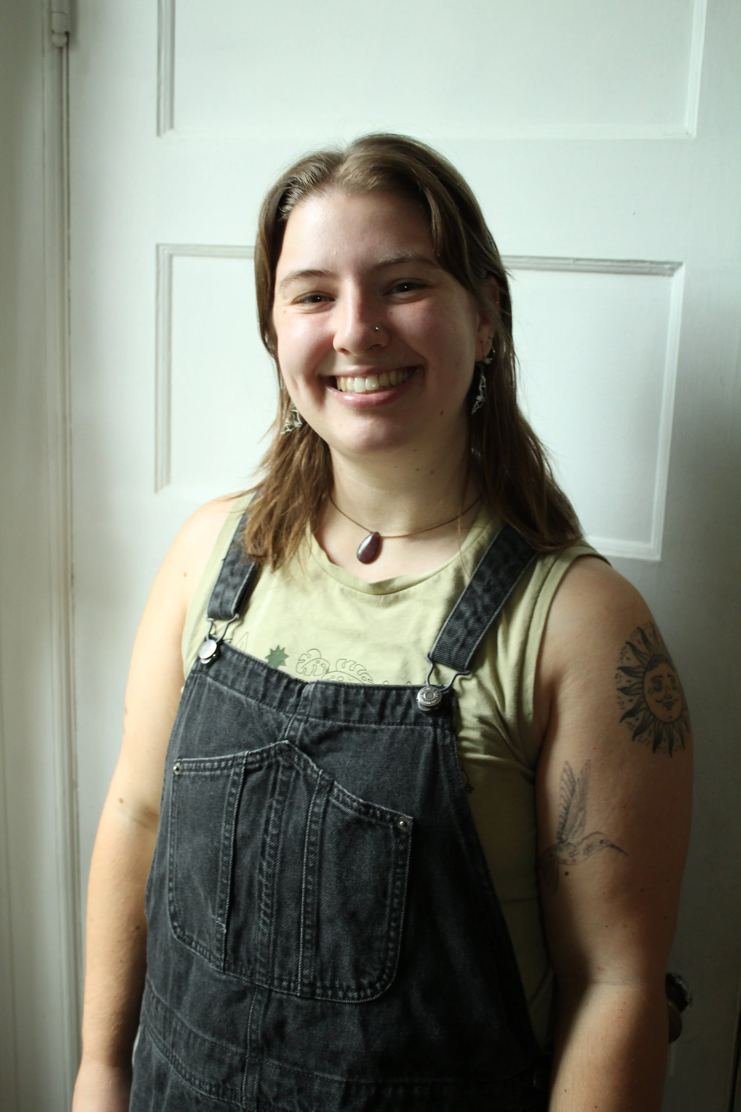 Home organizer Emilie with shoulder-length brown hair, wearing a light green tank top and black denim overalls, smiling in front of a white door.