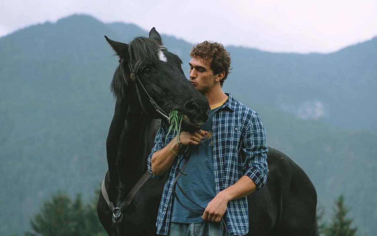 Un giovane uomo con capelli riccioli fa un gesto affettuoso vicino a un cavallo nero con una macchia bianca sulla fronte, tenendo un rametto di erba in bocca.