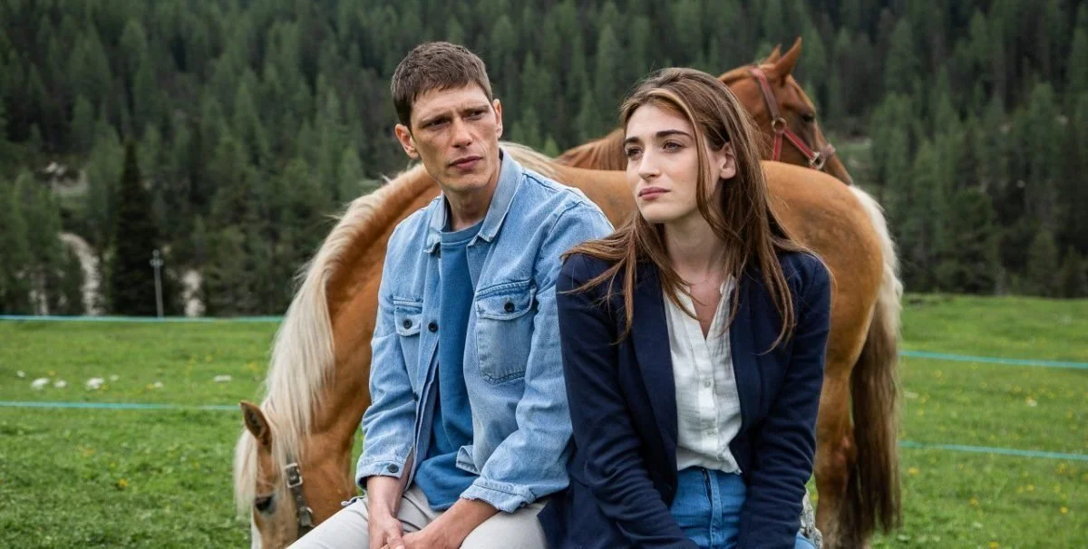 Un uomo e una donna seduti su una staccionata in un prato, con un cavallo sullo sfondo.