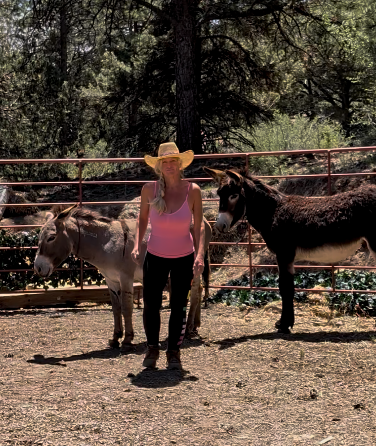 A woman wearing a cowboy hat, pink tank top, black pants, and boots standing between two donkeys in an outdoor enclosed area with trees and a metal fence.