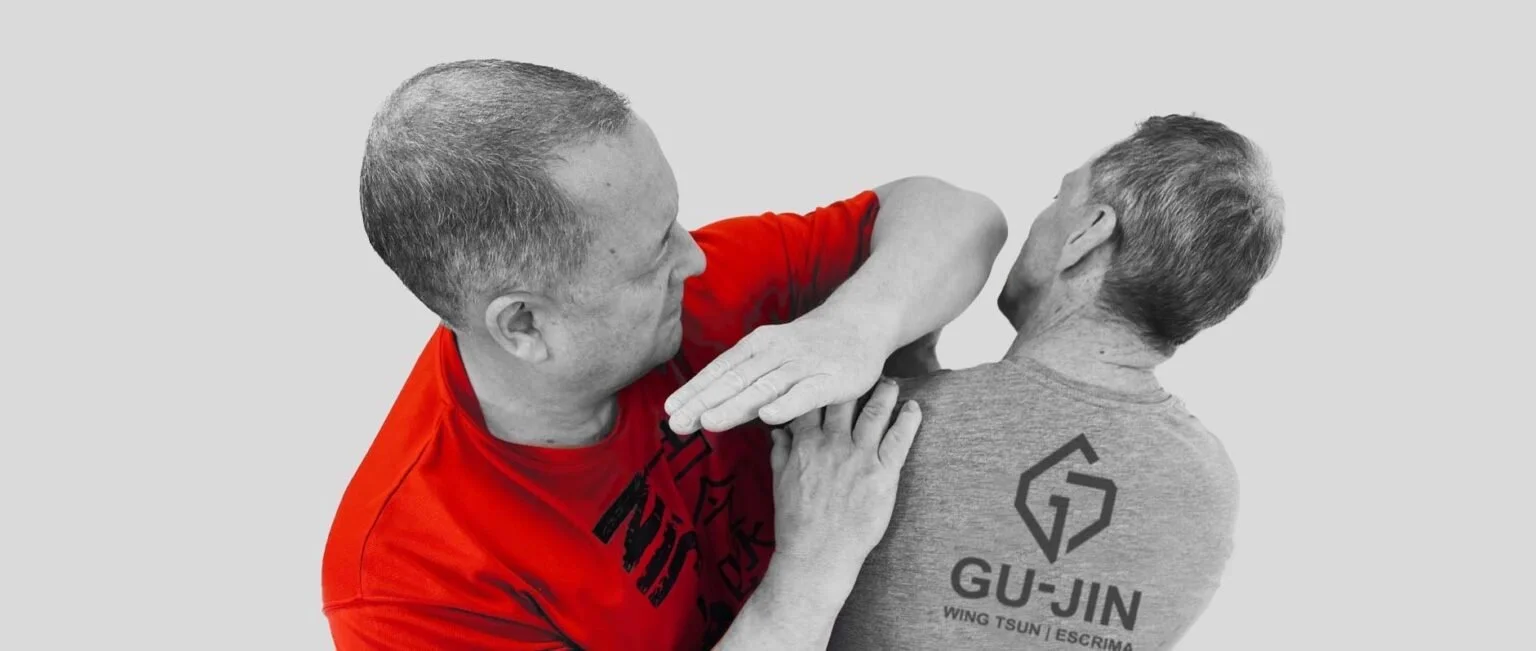 Two men practicing Wing Tsun martial arts; one in a red shirt demonstrating a defensive move against the other in a gray shirt with GU-JIN logo.