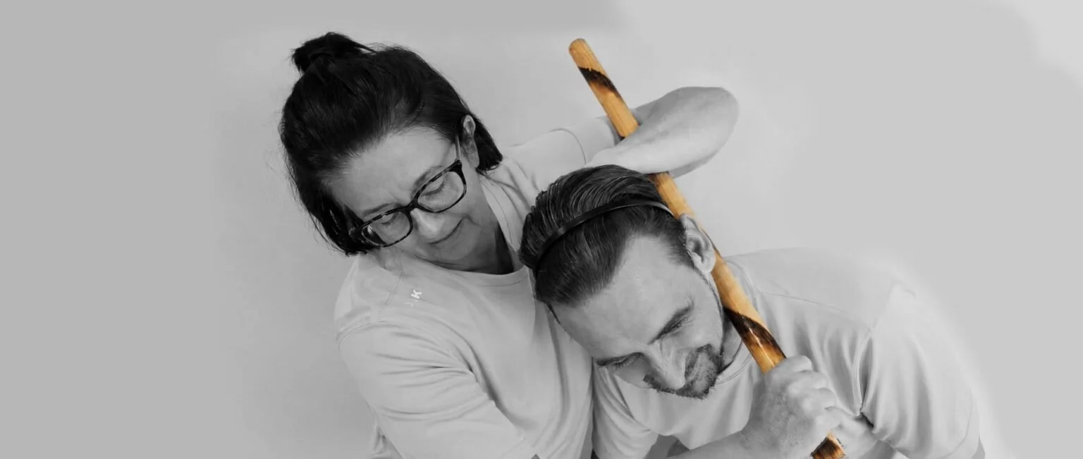 Two people, a woman with glasses and a man with a beard, engaged in a playful rock-paper-scissors game, each holding a wooden stick.