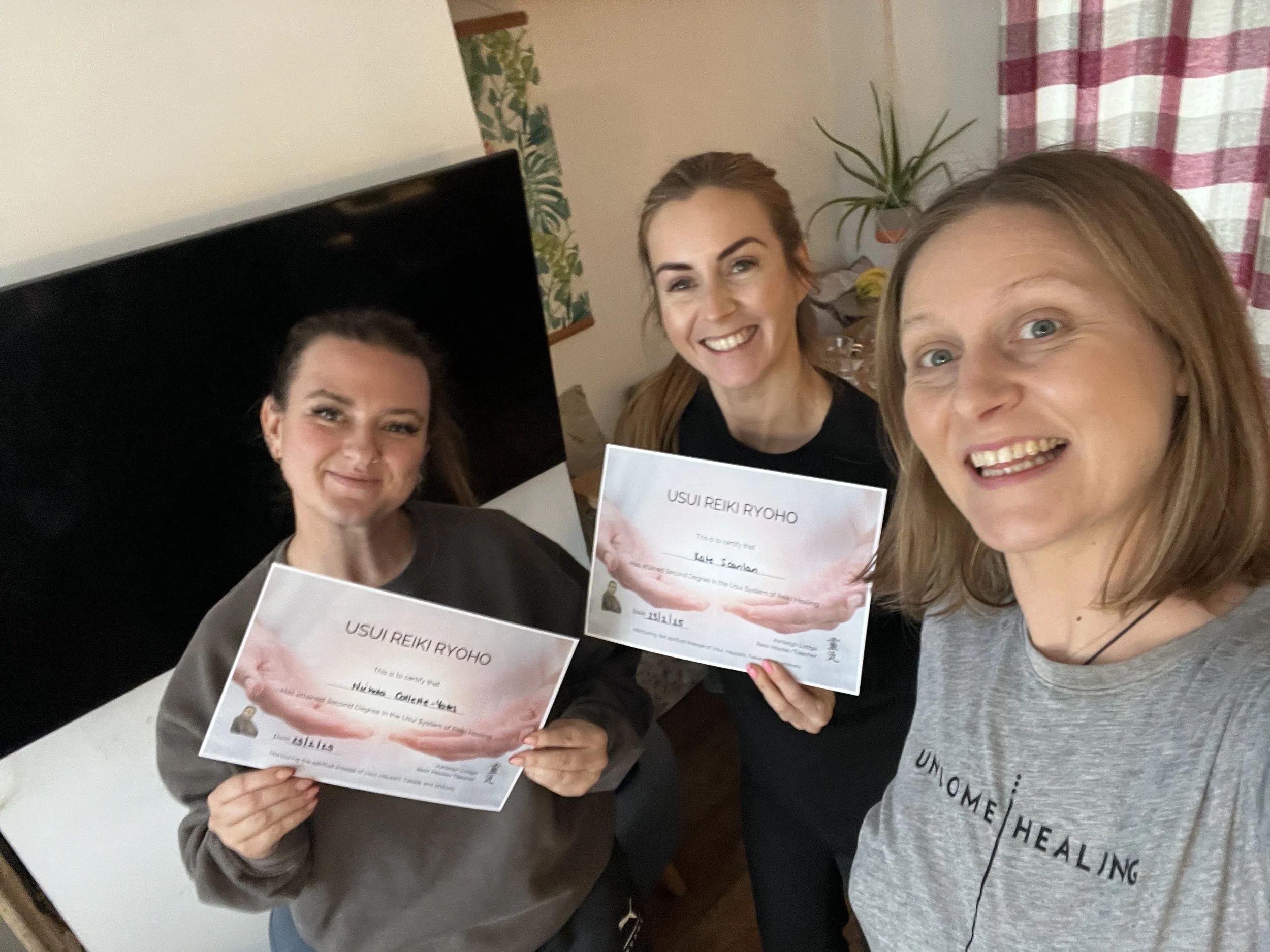 Three women smiling, holding certificates for Usui Reiki Ryoho, indoors, with a plant and a curtain in the background.