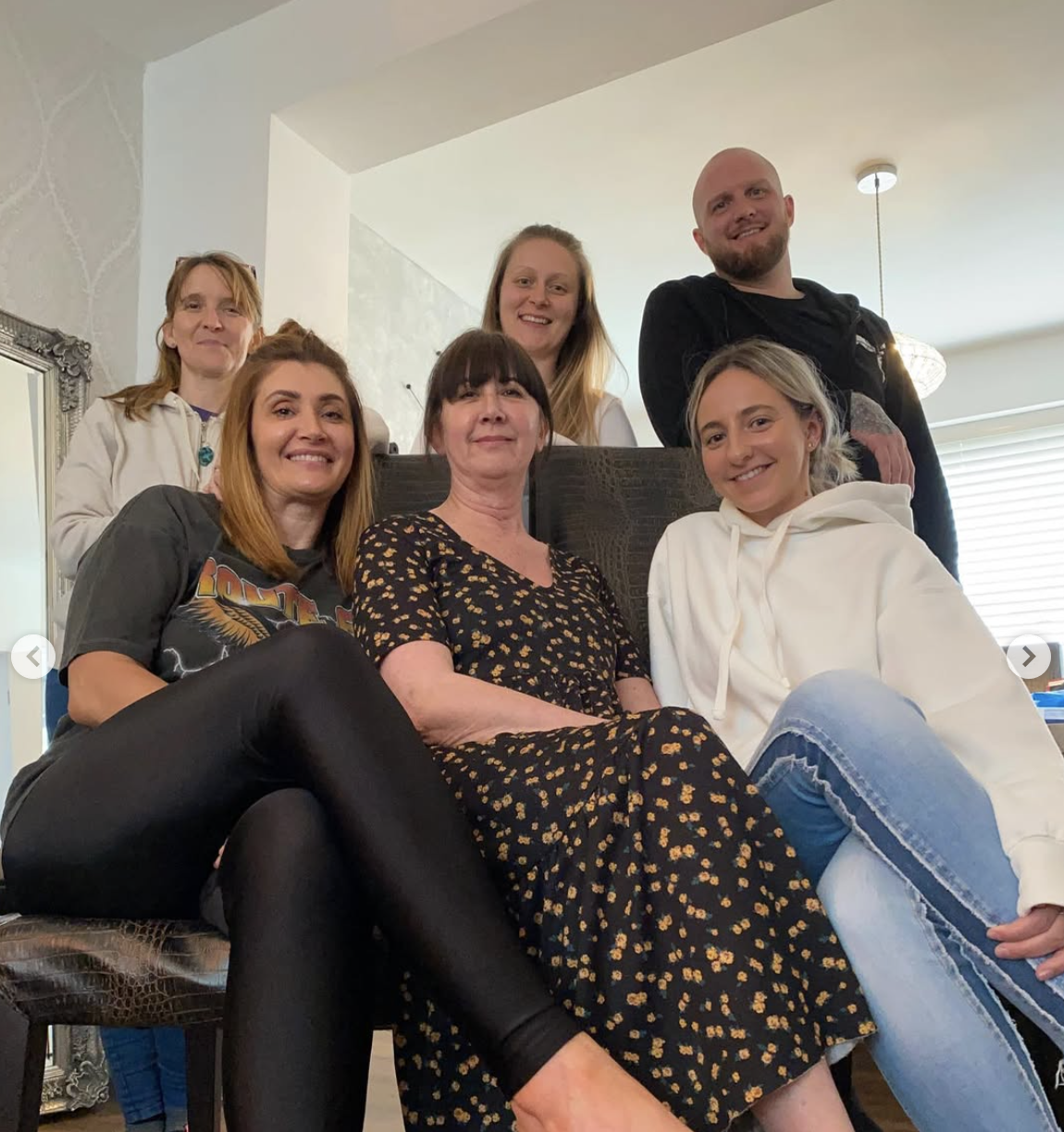 Six people gathered indoors, sitting and standing around on a living room, posing on a Reiki course. They are smiling at the camera, with various casual and stylish clothing, in a cozy setting.