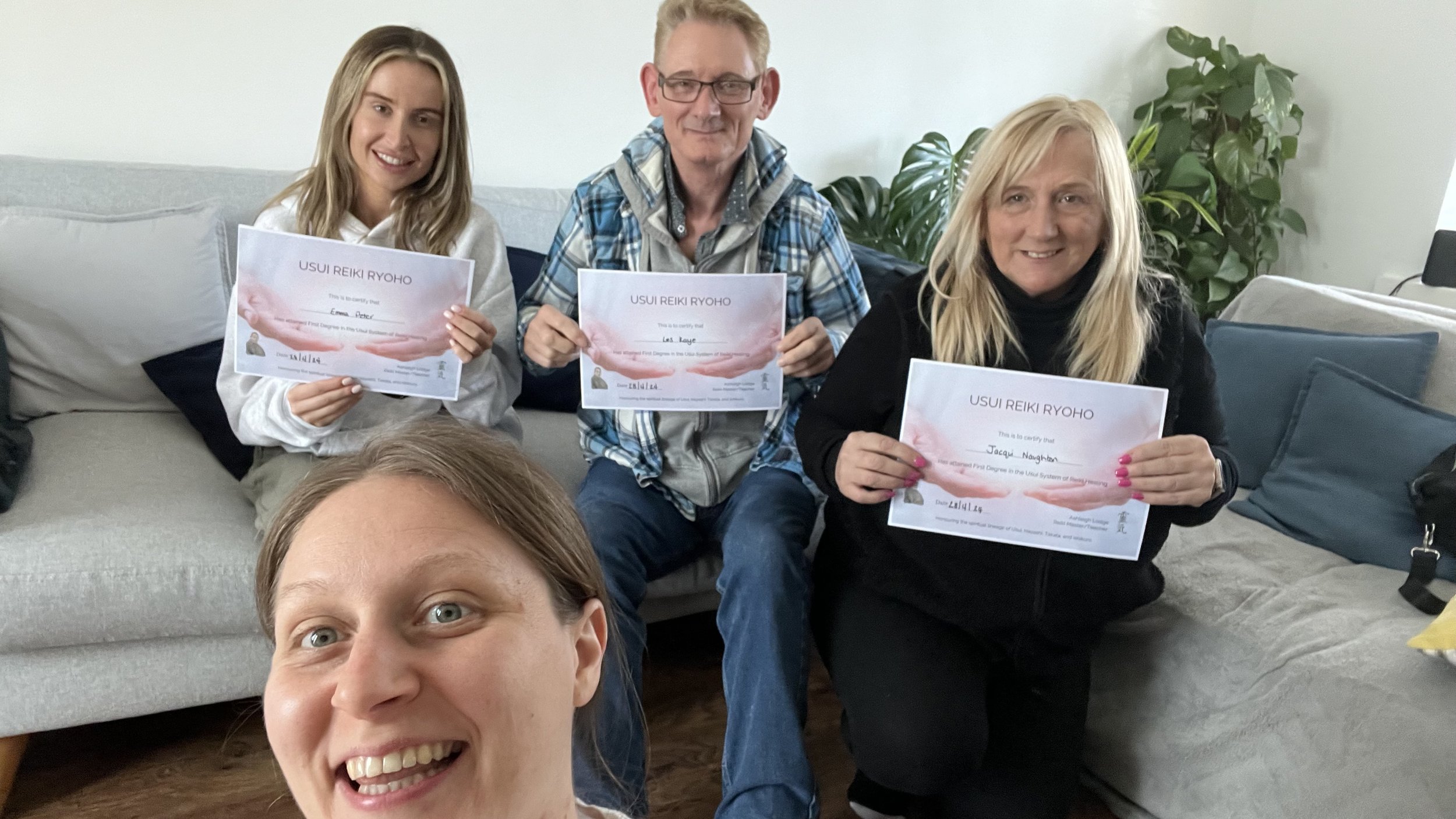 Three people sitting on a couch and holding Reiki certificates, with a large leafy plant in the background. One person is partially visible at the bottom of the image, taking a selfie.