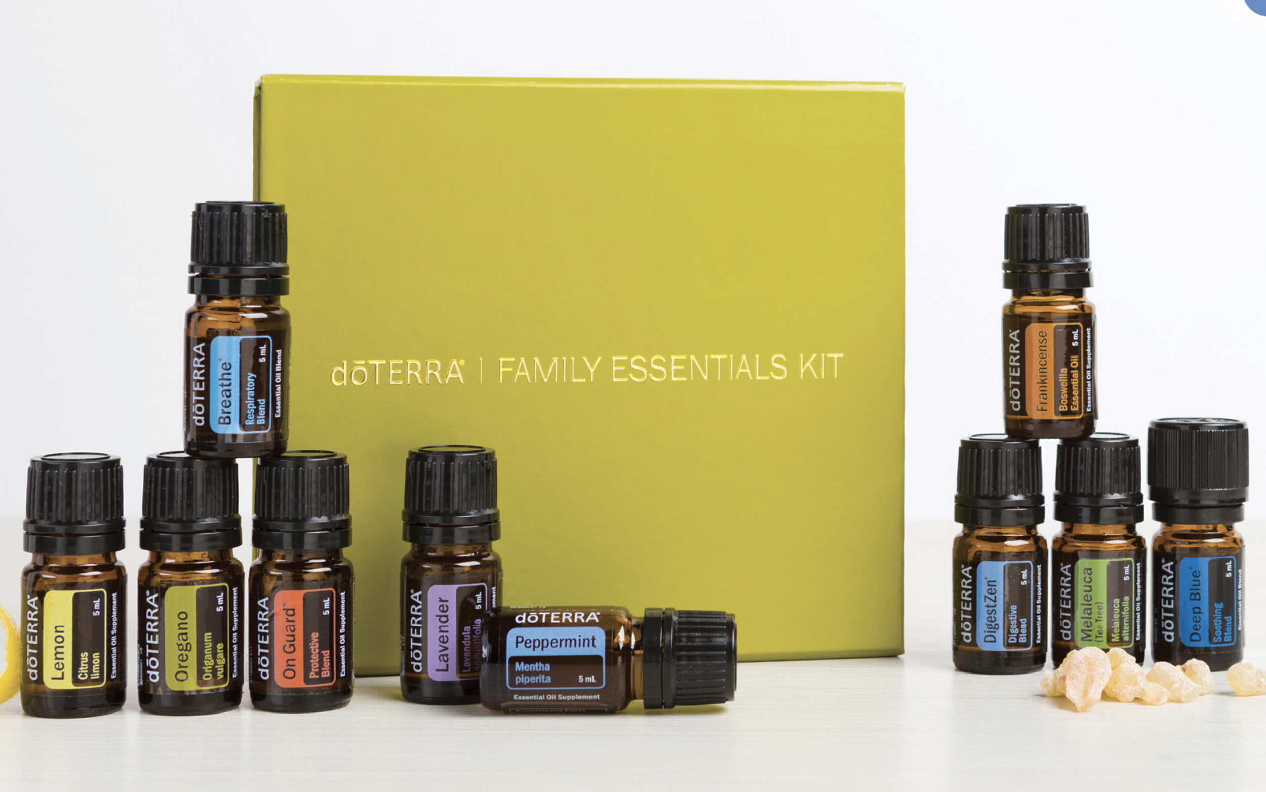 Collection of dōTERRA essential oil bottles arranged in front of a bright yellow box labeled 'dōTERRA Family Essentials Kit', with some essential oils visible such as Peppermint and Lavender, and small chunks of white rock sugar on the side.